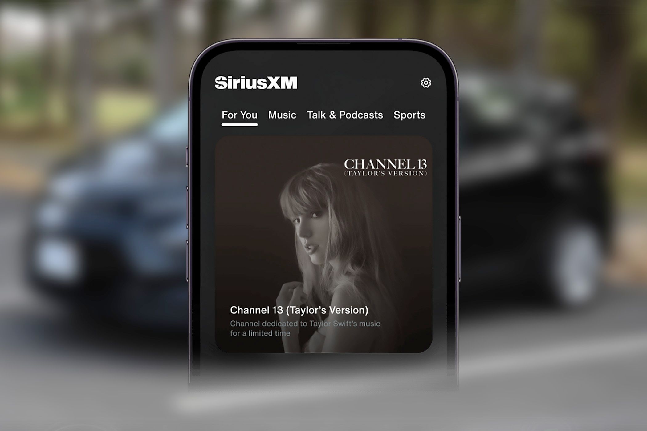 A phone playing SiriusXM Channel 13 with a car in the background.