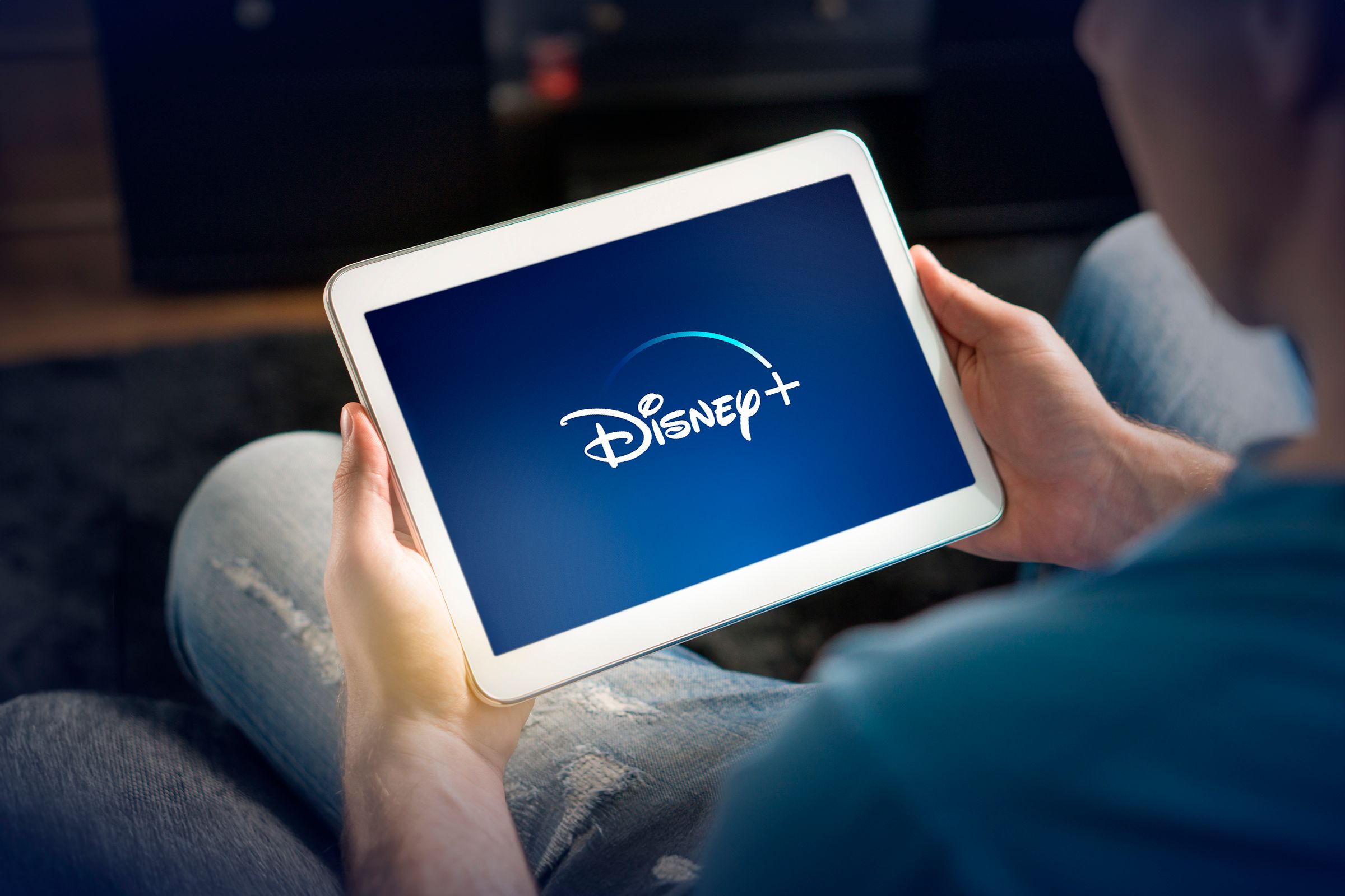 A man on a couch holding a white tablet with Disney+ logo on it.