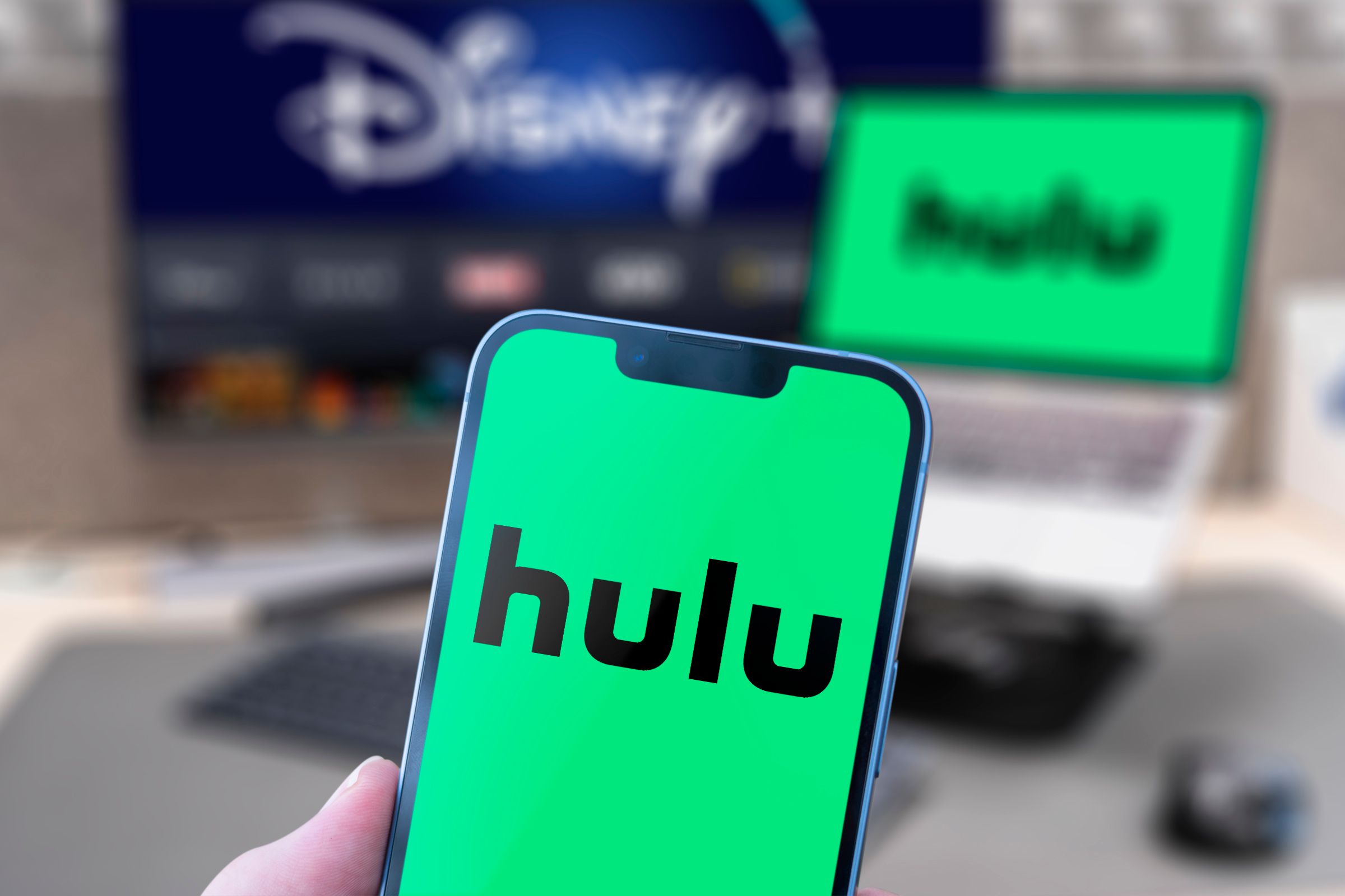 A phone with the Hulu logo and a monitor and laptop with the Disney Plus and Hulu logo blurred in the background.