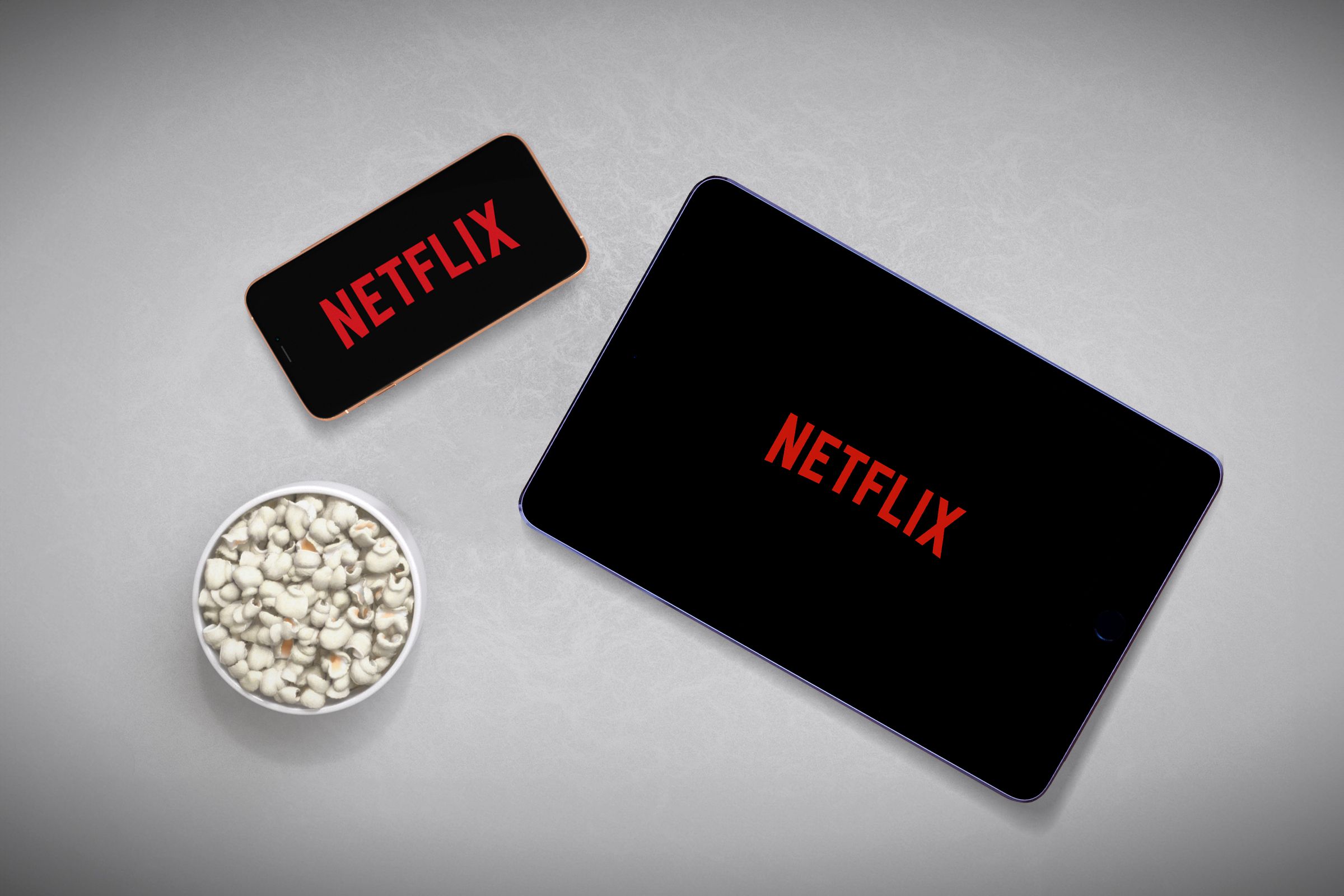A smartphone on the left and a tablet on the right, both with the Netflix logo on the screen, and a bowl of popcorn in the bottom left corner