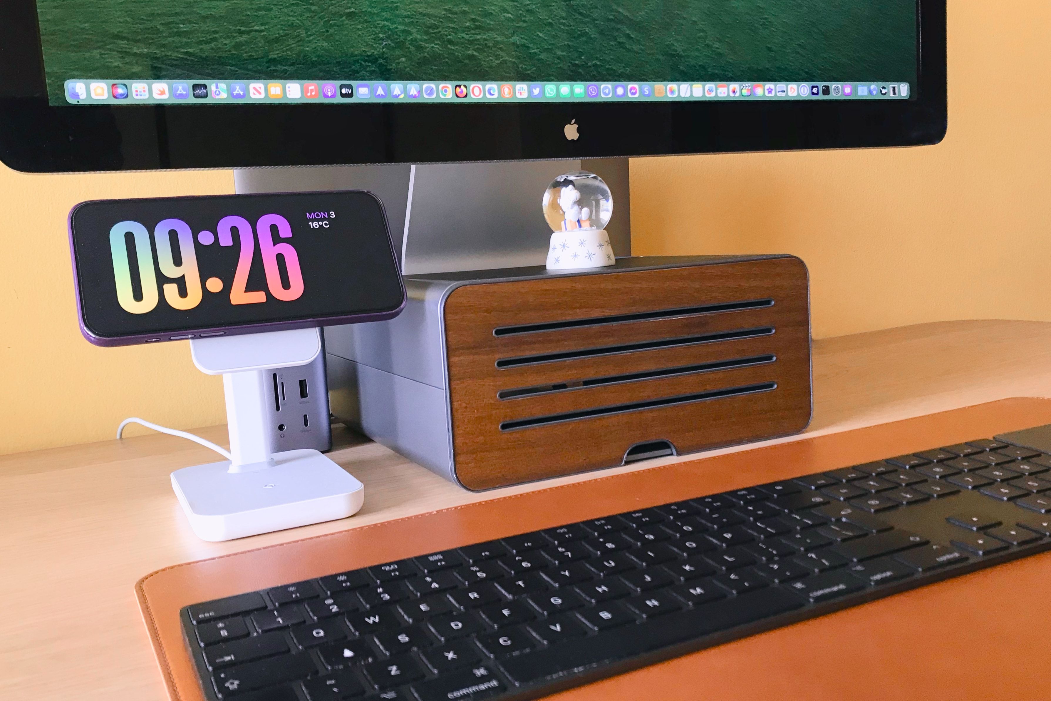 An iPhone in StandBy Mode on a MagSafe charging stand sitting on a wooden desk with a keyboard and monitor