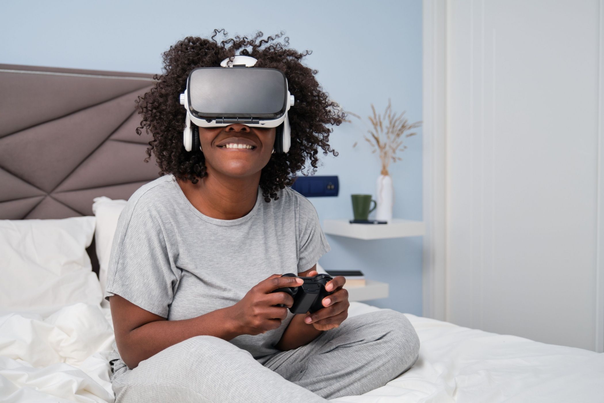 A young woman playing video games with a VR headset sitting on bed in the morning.