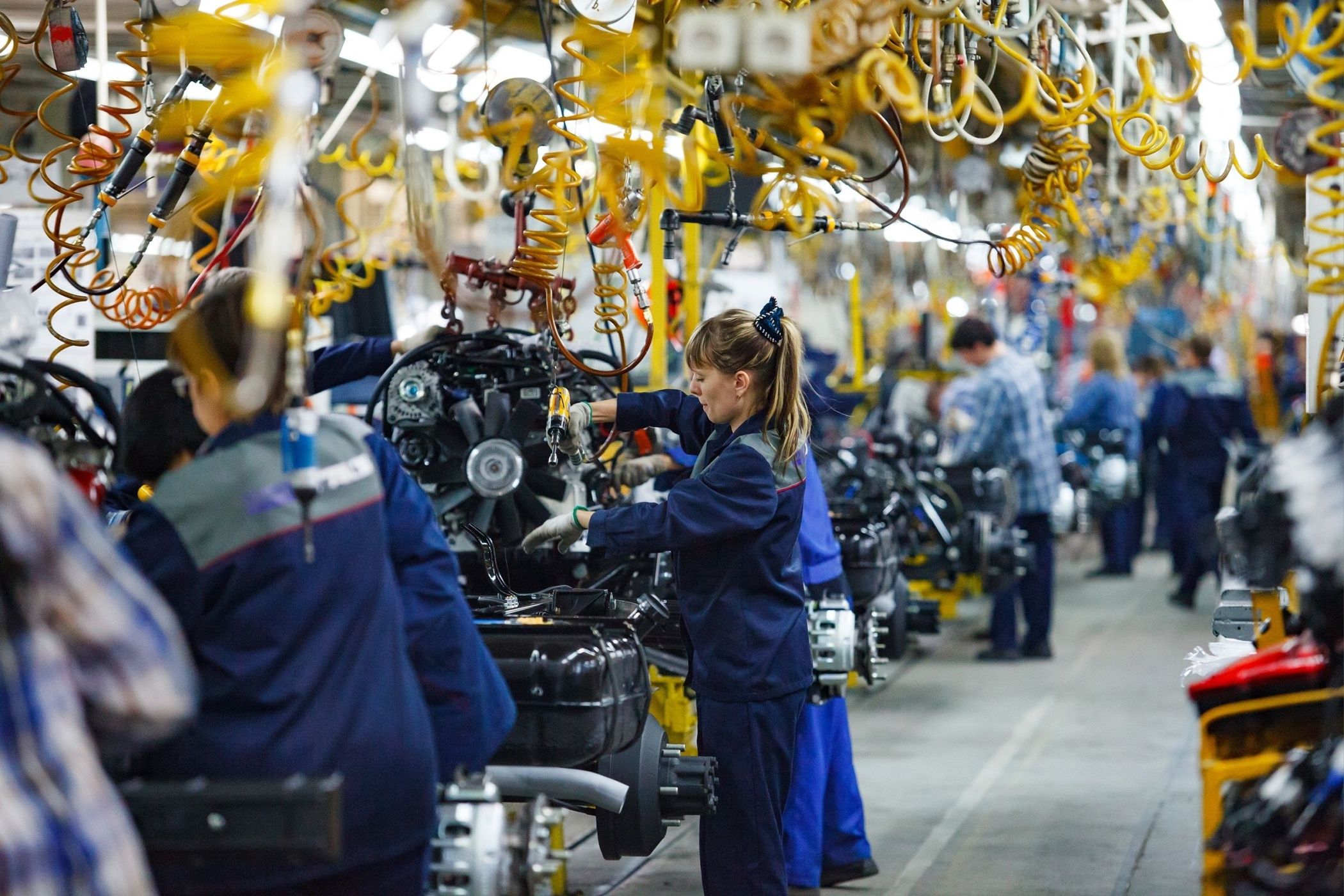 car production plant. People work on assembling line. Young woman in focus only.