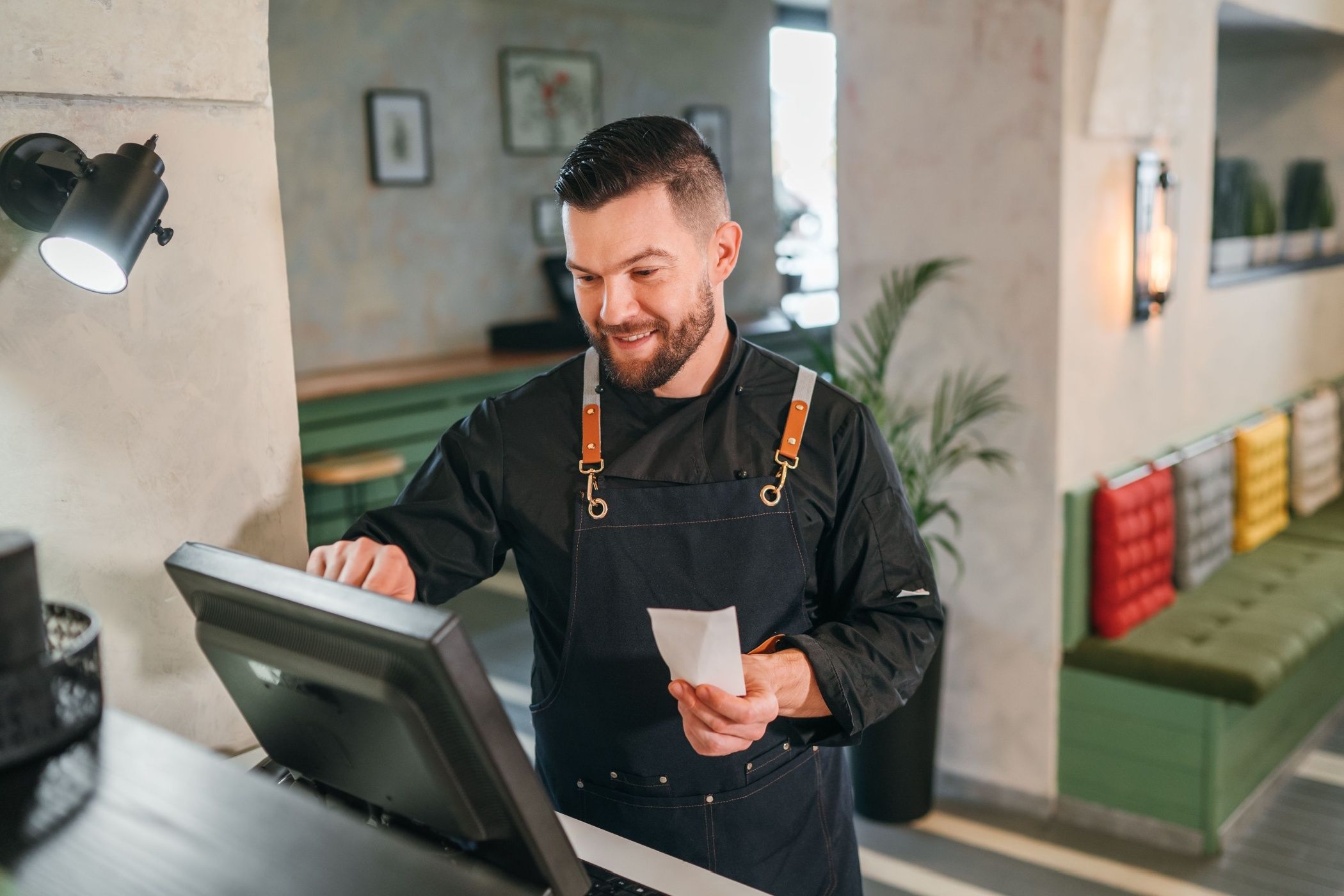 Stylish bearded smiling waiter dressed black uniform processing customer orders using point of sale order terminal system touch screen.