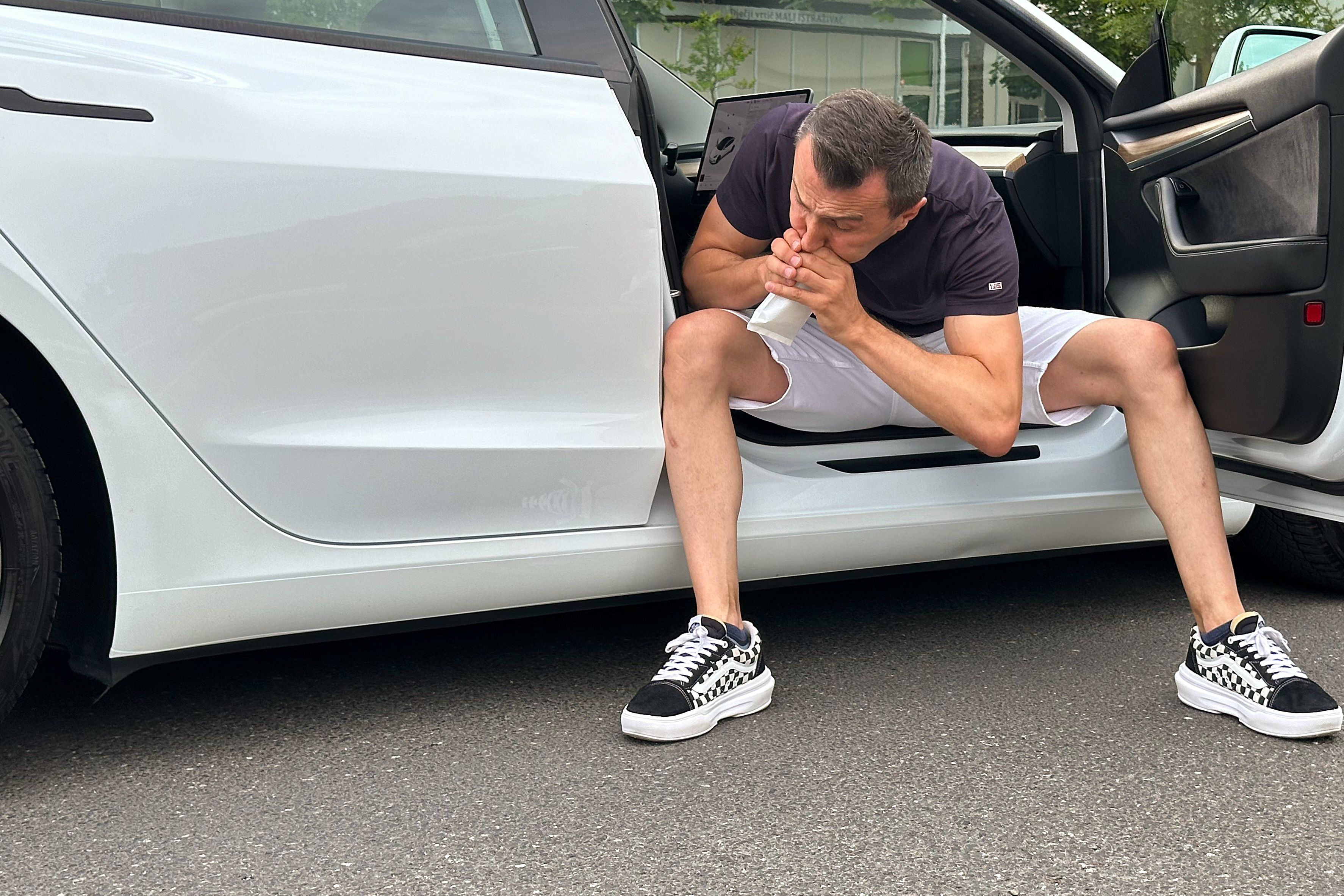 A white Tesla with its passenger door open and a passenger sitting with their legs outside and vomiting into a white plastic bag.