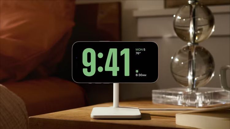 iPhone on wireless charging stand in StandBy Mode, showing time, weather, and current alarm.