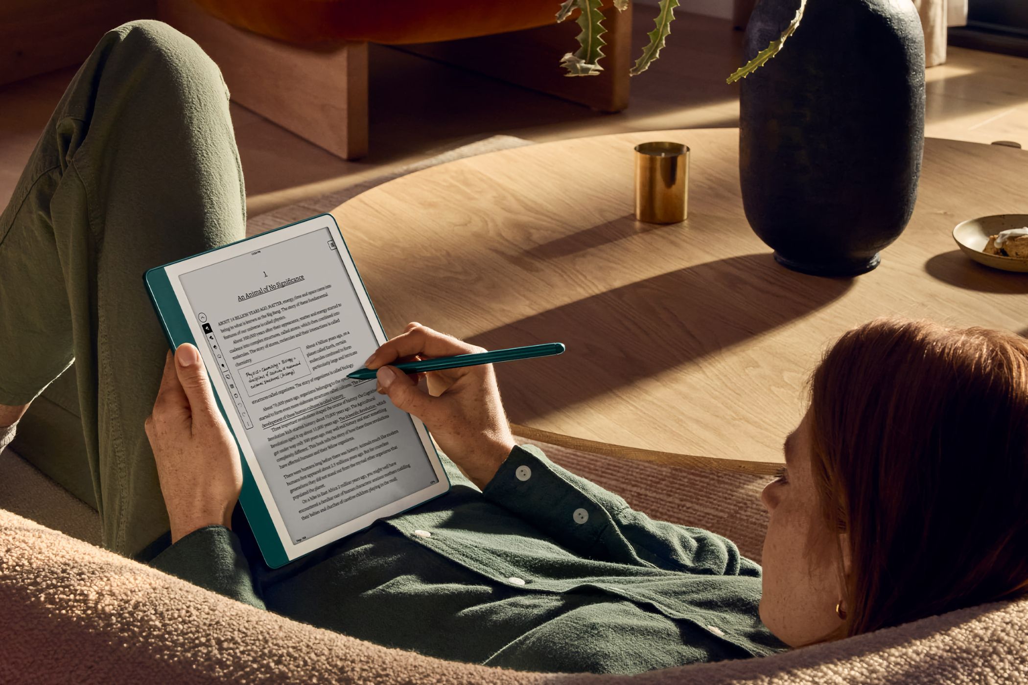 Woman lying on a couch, using Amazon's Kindle Scribble reader with a stylus to add inline notes.