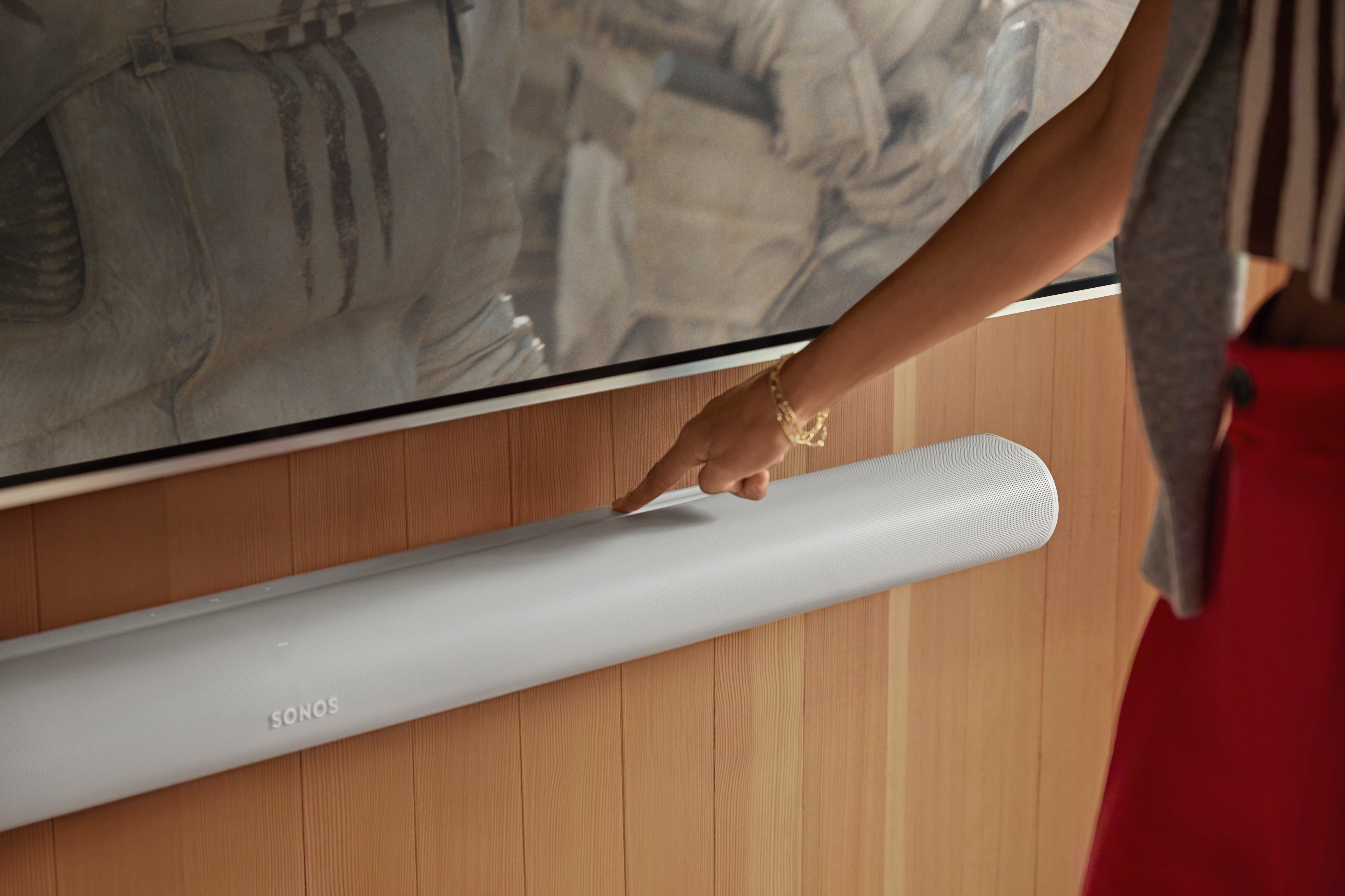 Female hand touching a Sonos Arc Ultra soundbar below a huge TV set.