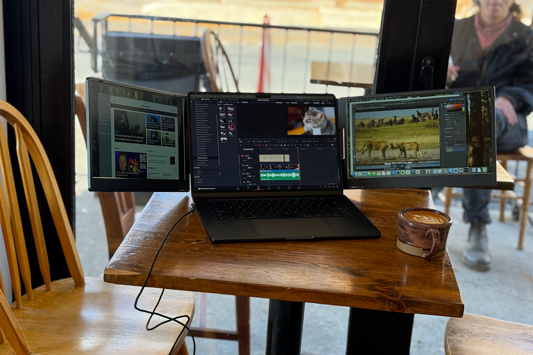 A MacBook Air sits near a window in a coffee shop displaying three windows including a browser, Resolve, and photo editing tool.