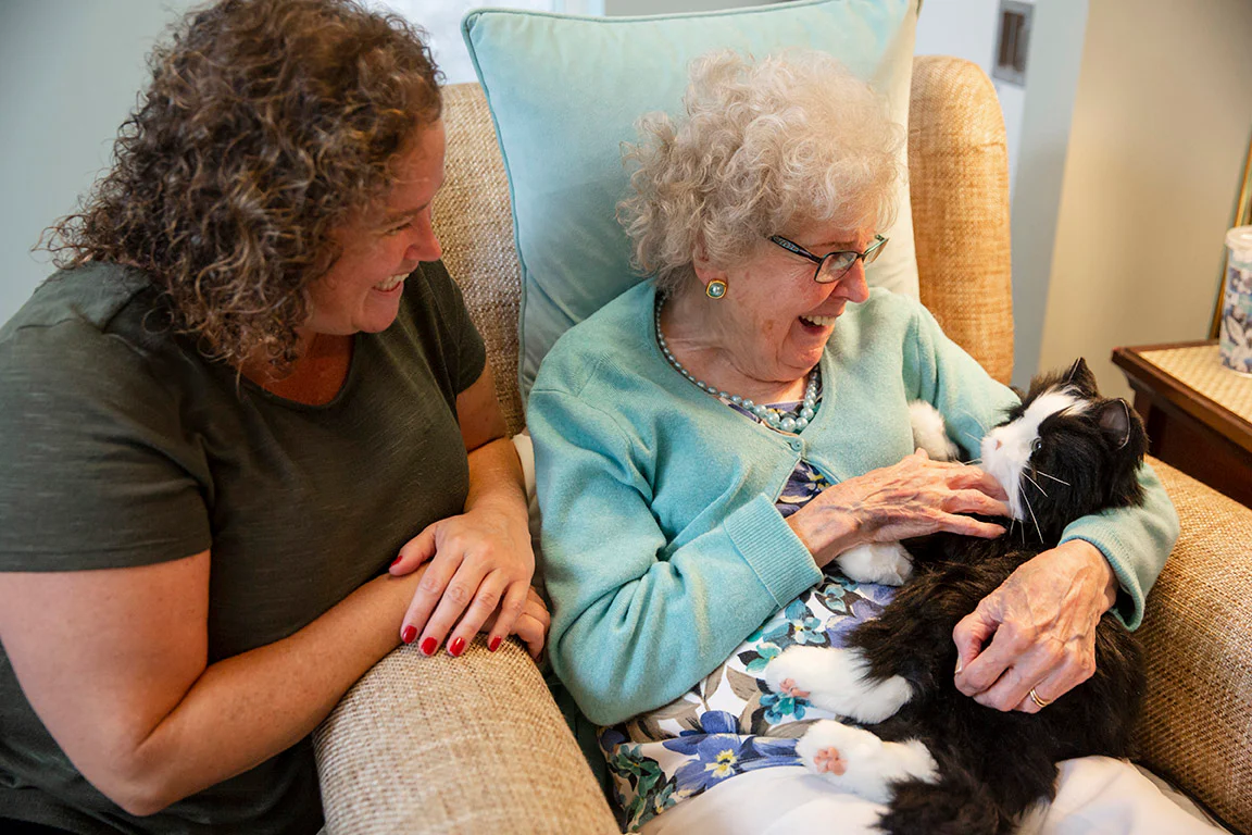 An elderly woman holding a robot cat while a middle-aged woman watches.