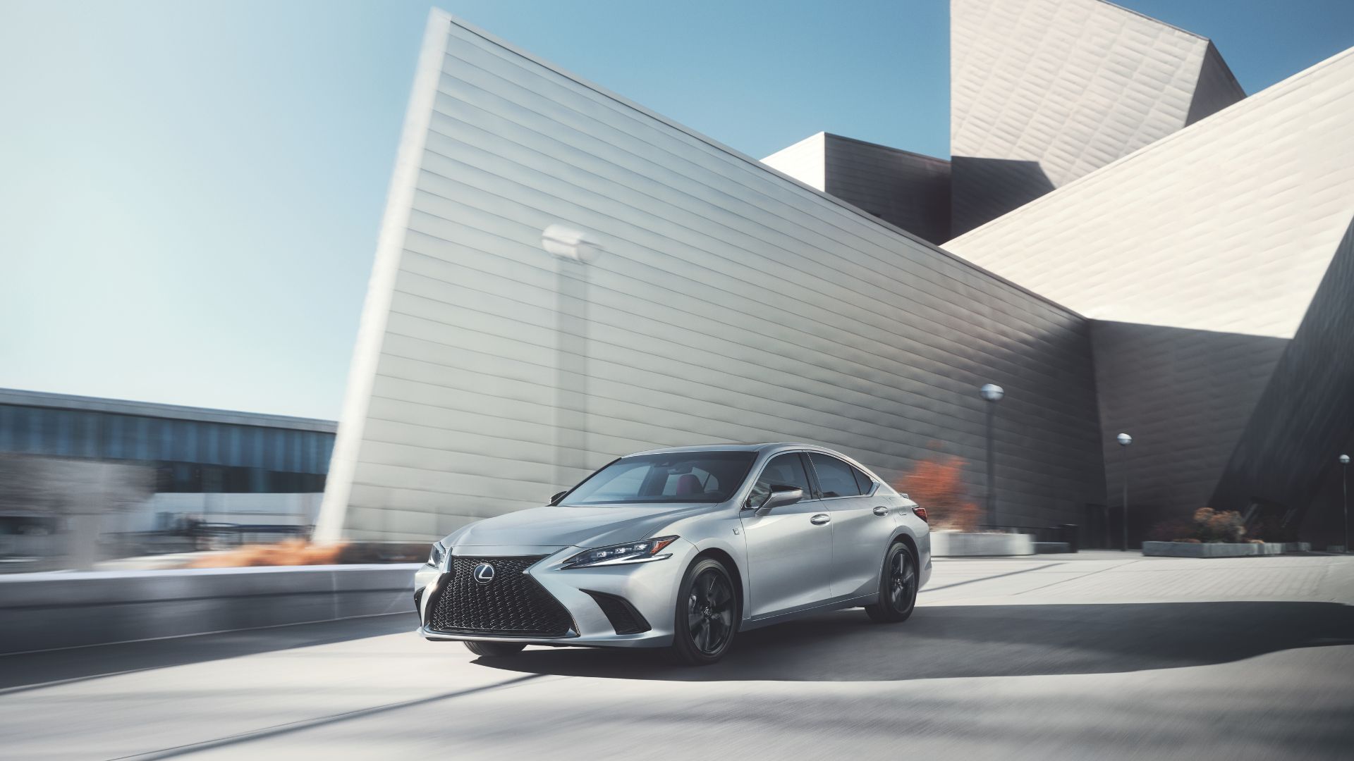 A silver 2023 Lexus ES 300h driving on a road in front of a modern building.