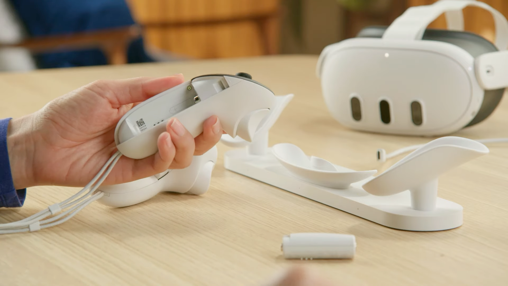 A hand holding a Meta Quest 3 Touch Plus controller with a rechargeable battery, and a Meta Quest 3 and charging dock in the background.