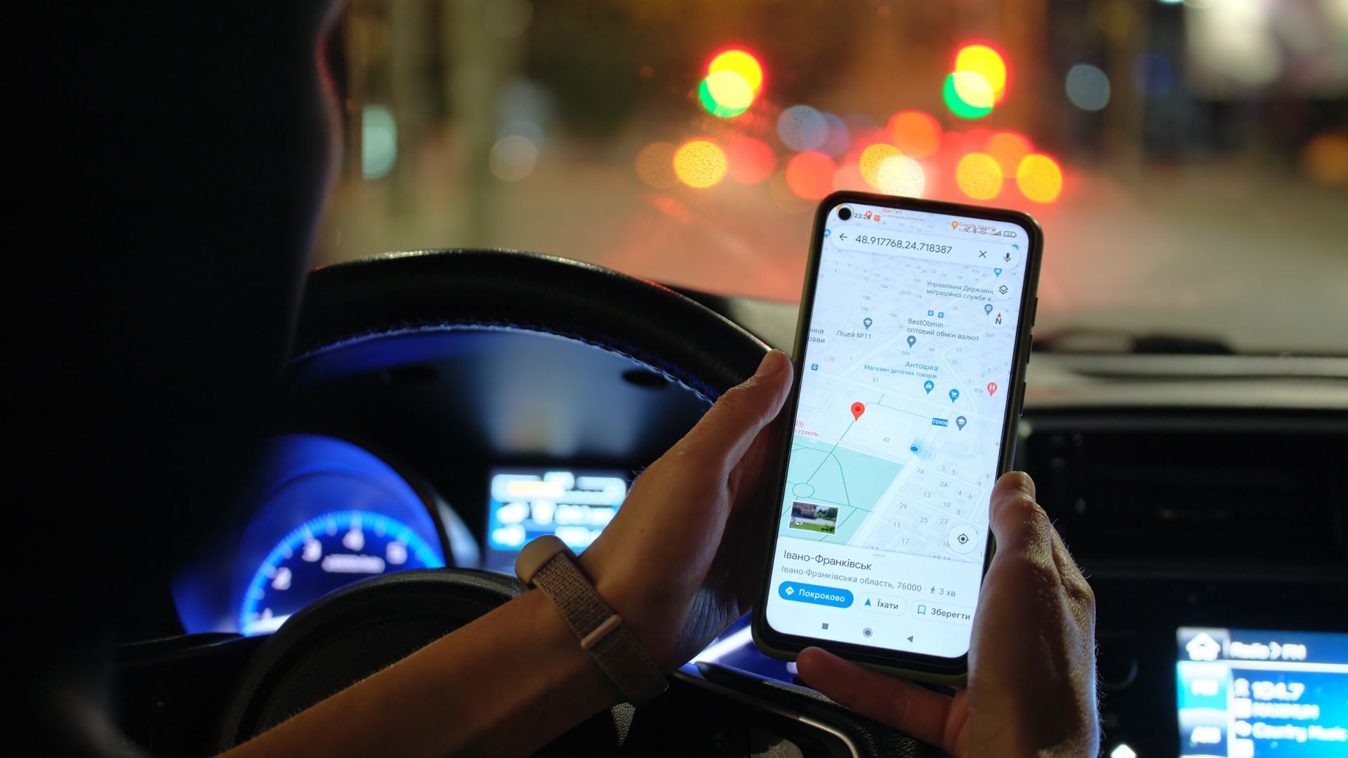 Close up of driver hands holding smartphone with Google Maps navigation app searching for direction with blurred city street lights in background at night.