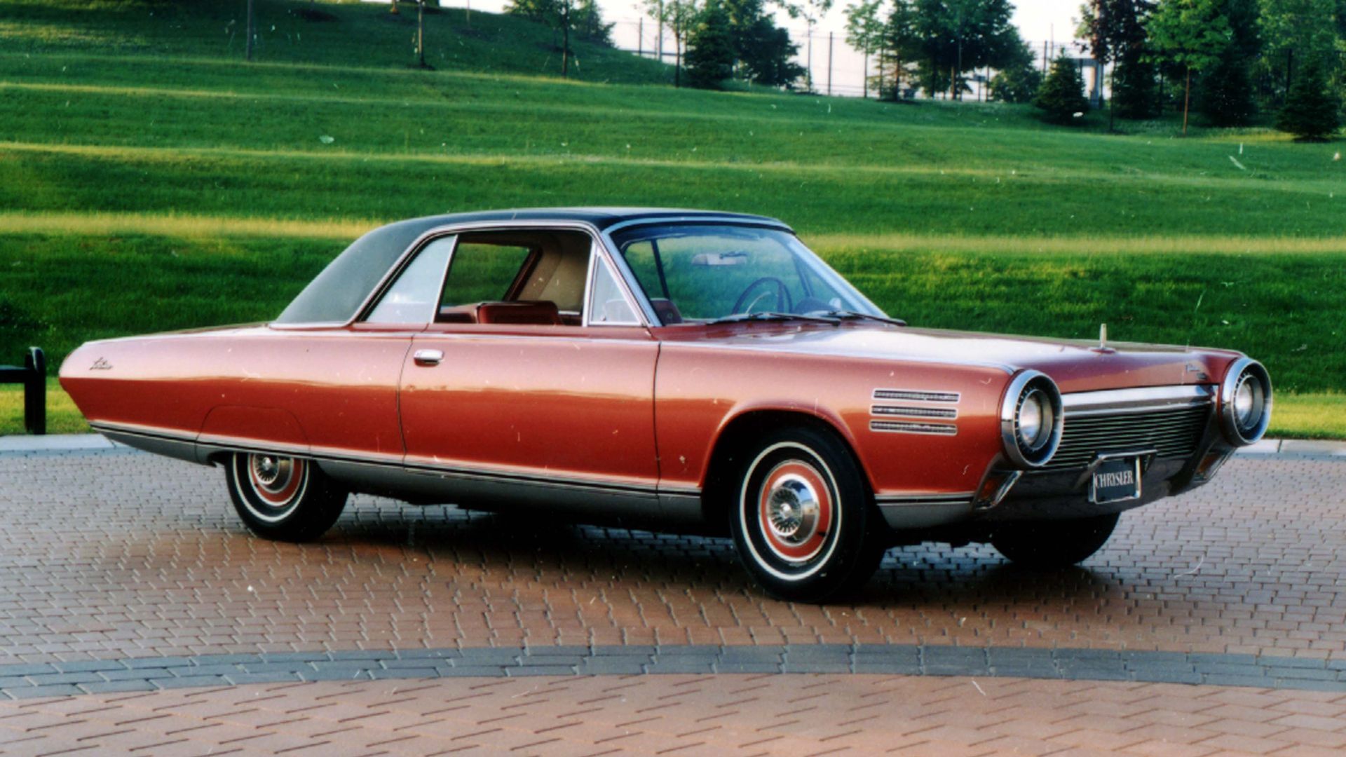 Side profile of an orange 1963 Chrysler Turbine Car parked on paving with grass in the background.