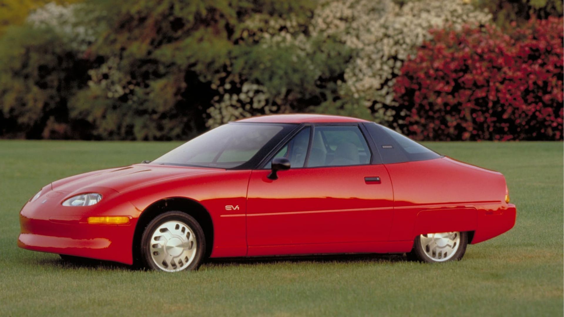 Side profile of a red 1996 General Motors EV1 parkerd on grass.