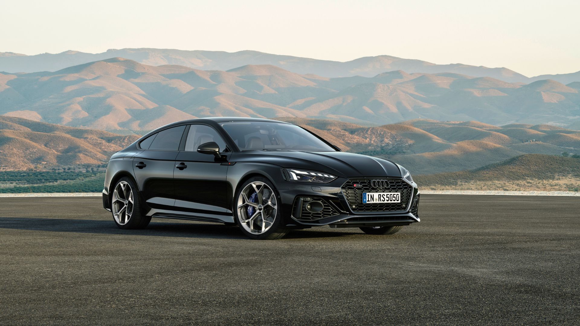 Full view of a black Audi RS5 Sportback parked on tarmac with mountains in the background.