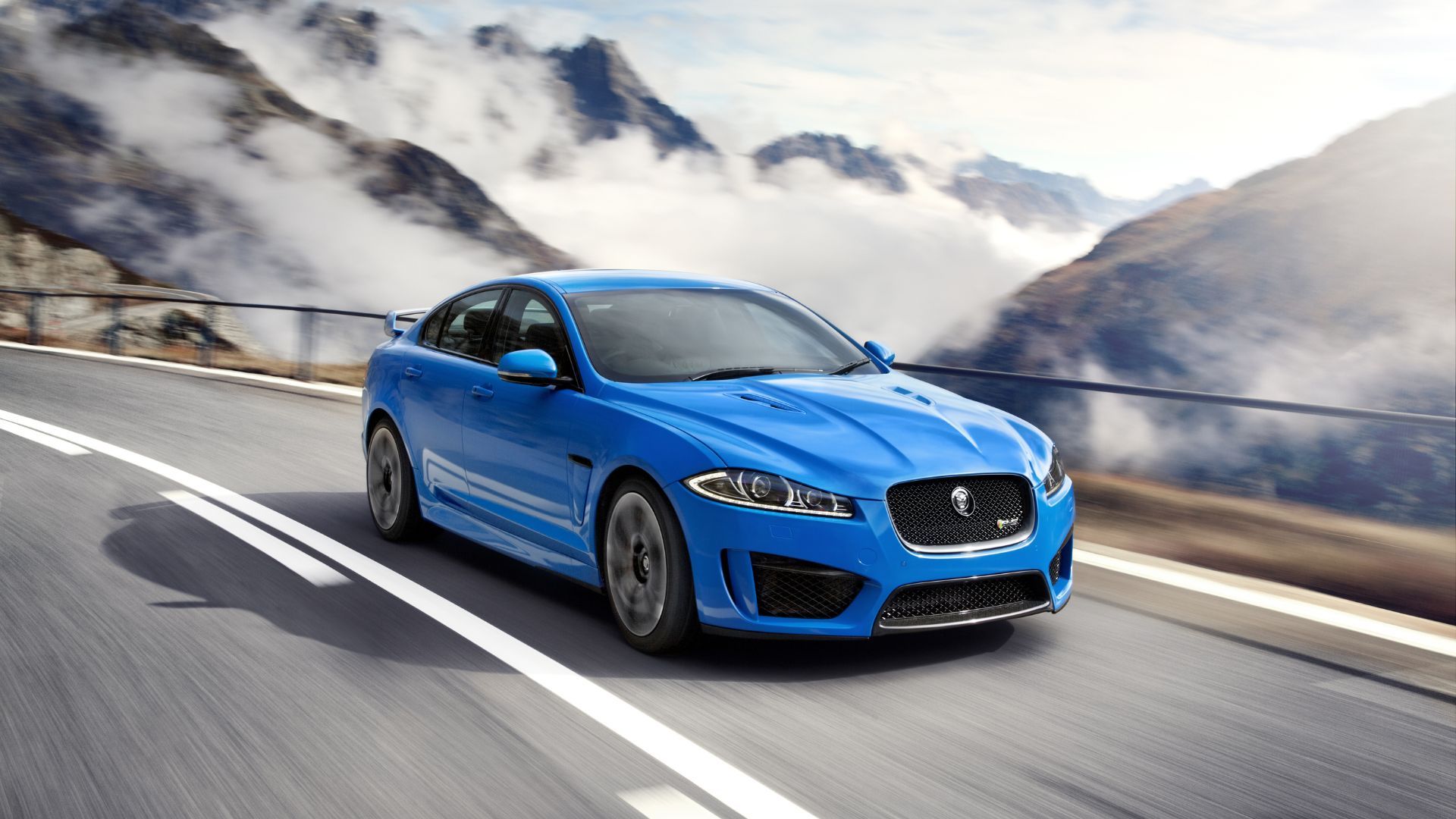 Full view of a blue 2015 Jaguar XFR-S driving on a winding country road with moutains in the background.