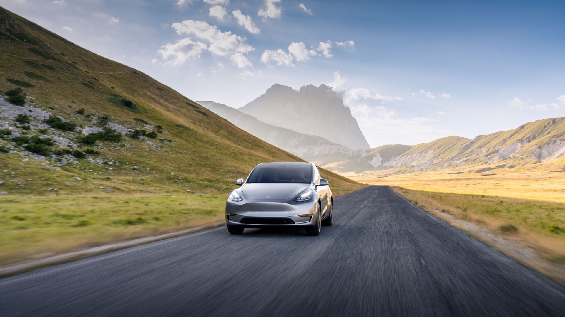 Silver Tesla Model Y driving on a country road with mountains in the background.