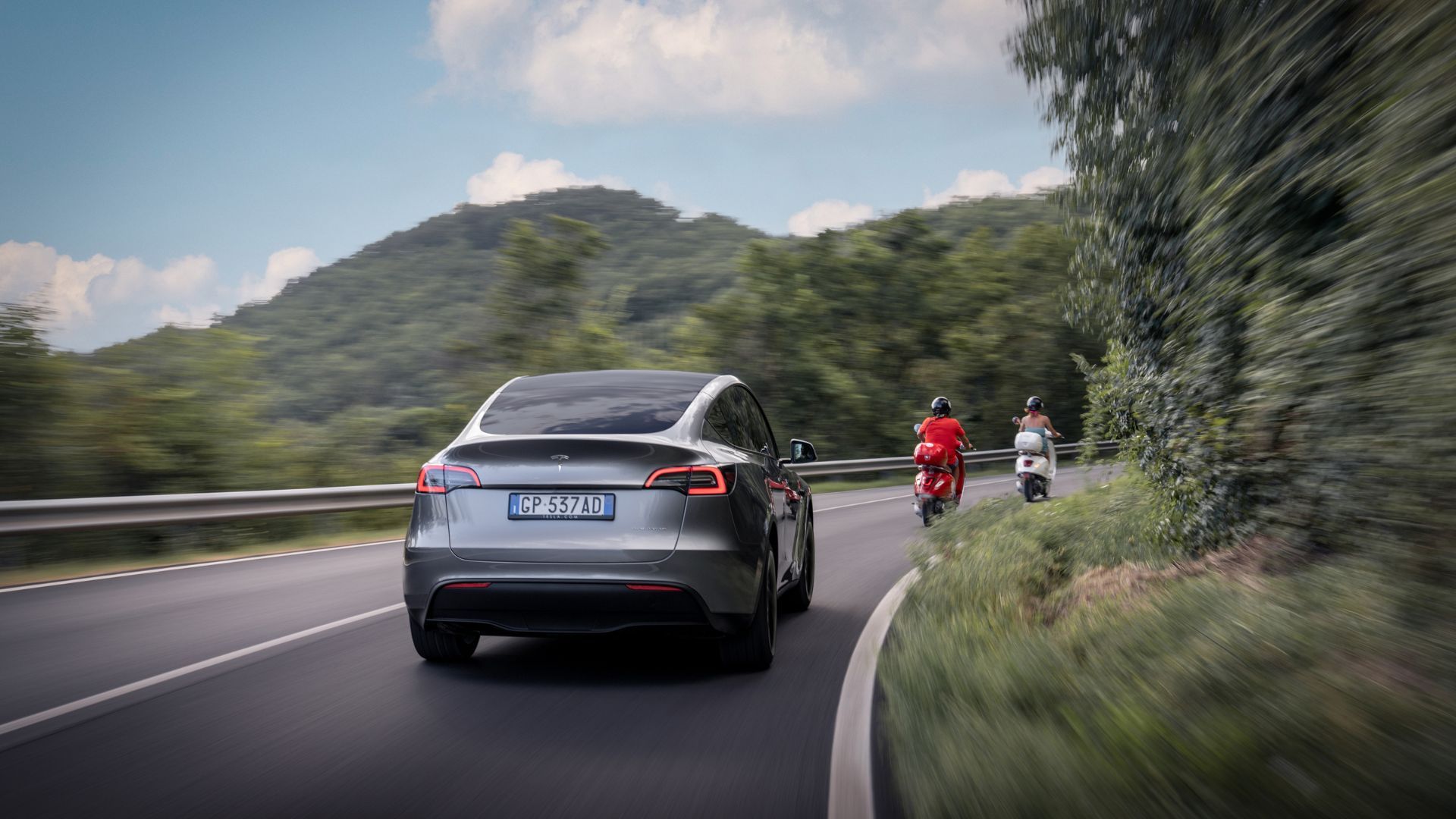 A silver Tesla Model Y driving on a winding country road following two motorcyclists.