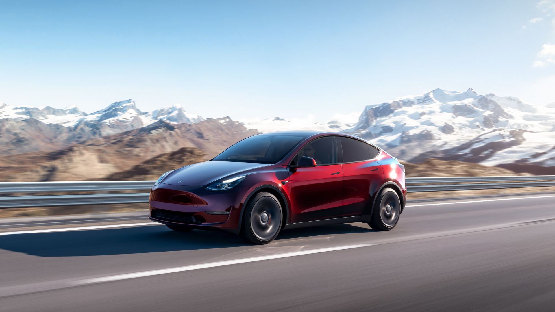 Red Tesla Model Y driving on a country road with snowcapped mountains in the background.