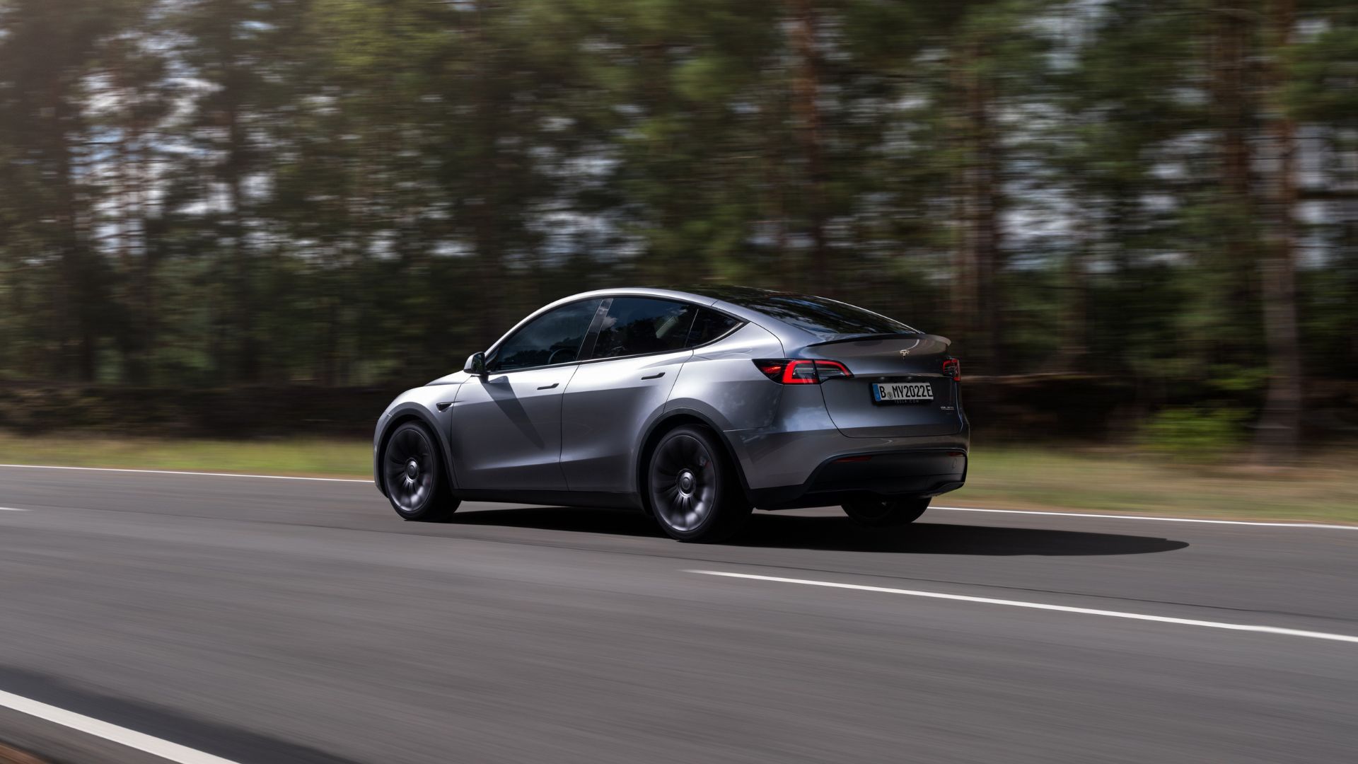 A silver Tesla Model Y driving on a country road through a forest.