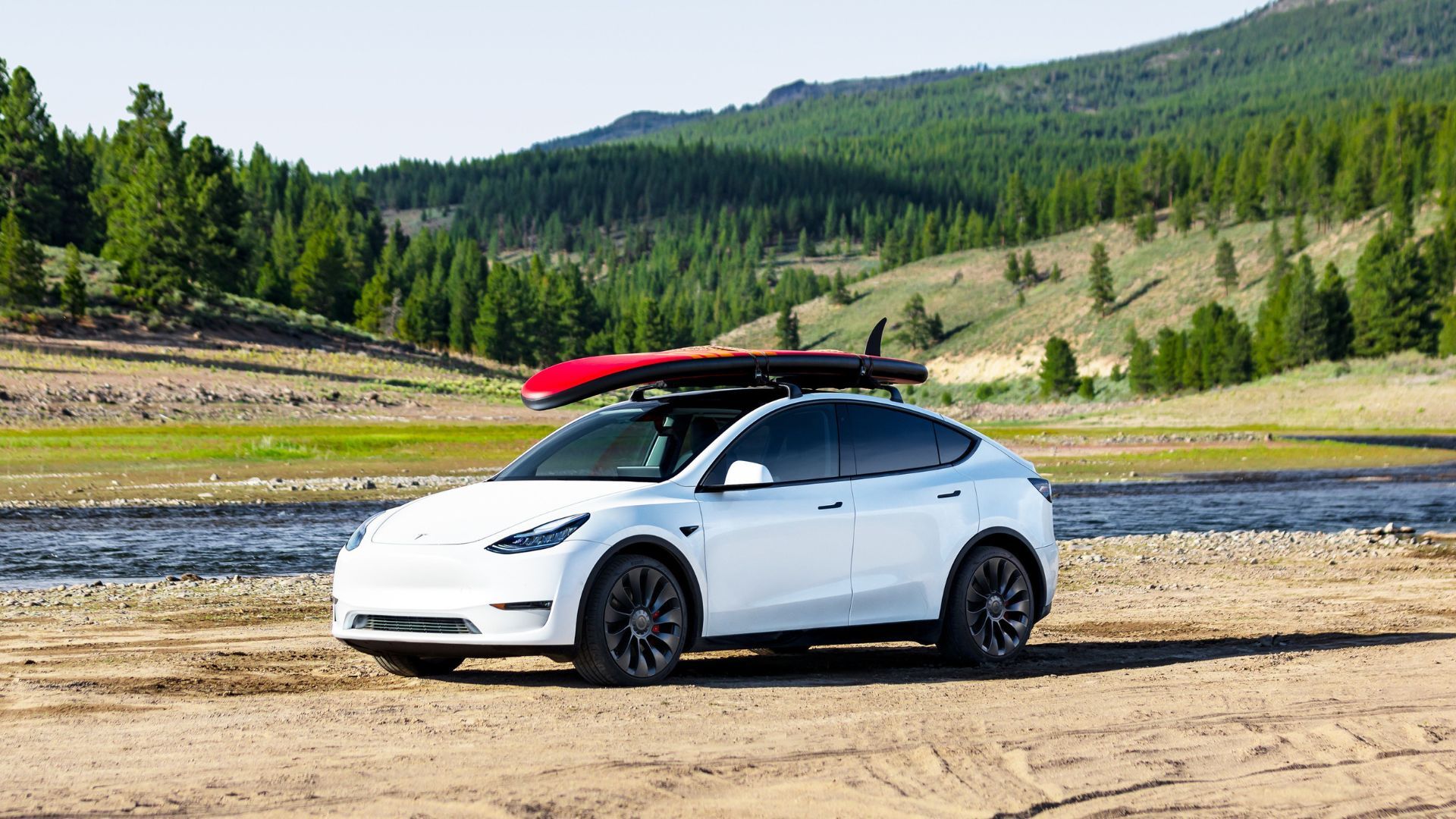 White Tesla Model Y with a paddle board on its roof parked on sand.