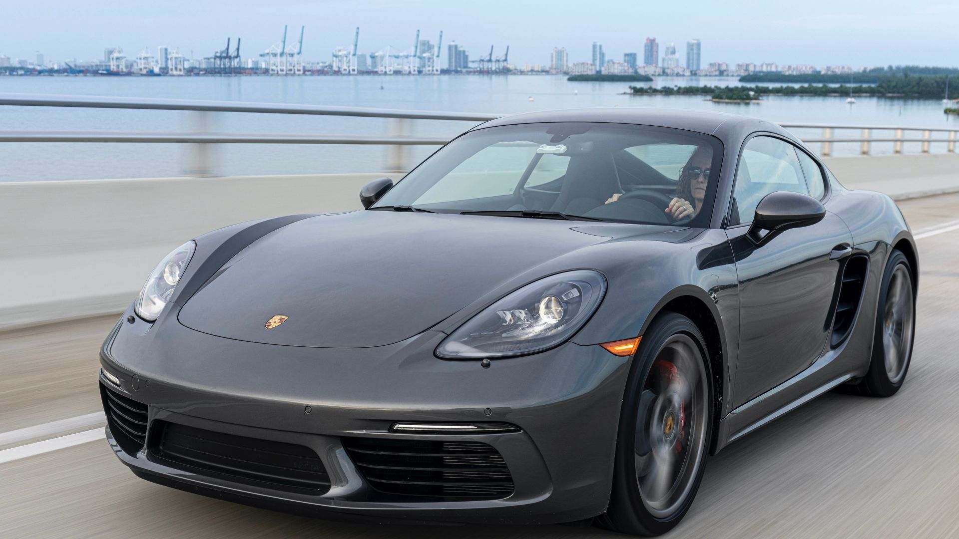 Full view of gray POrsche 718 Cayman driving on a bridge with Miami in the background.