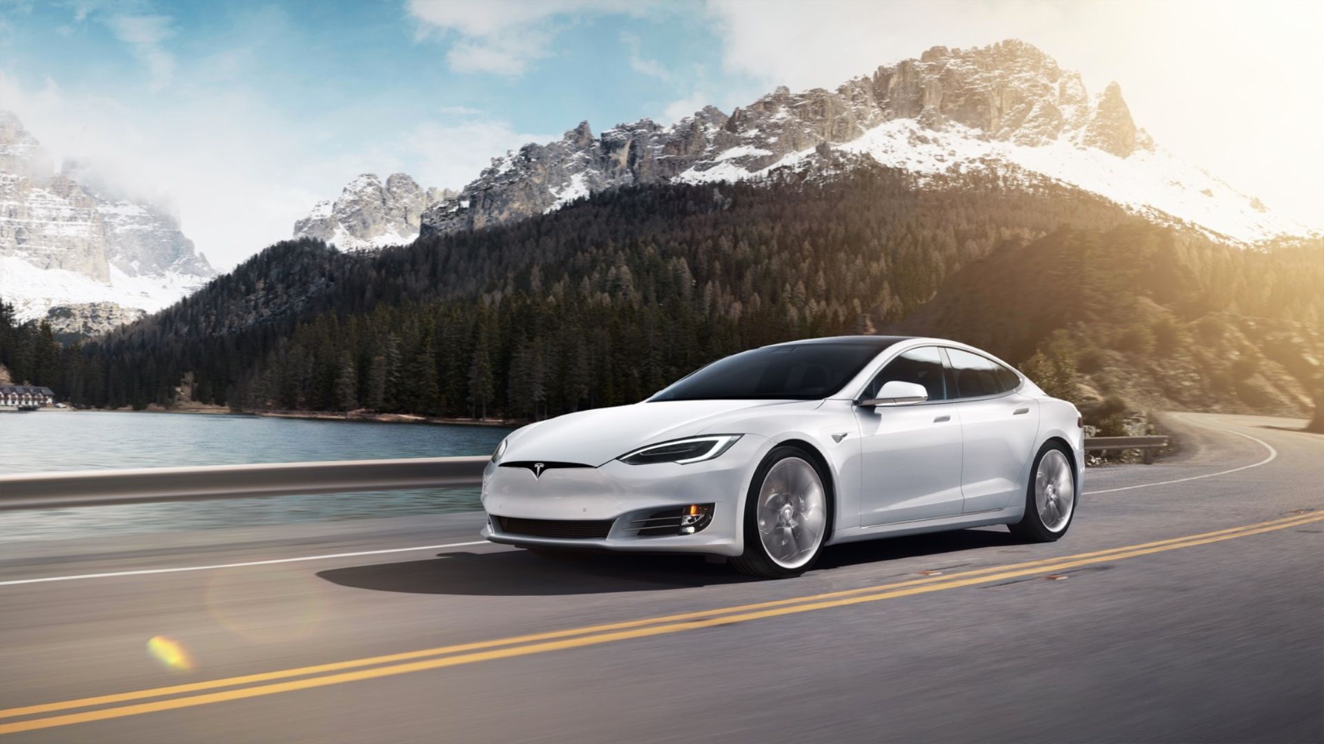 Full view of a white 2016 Tesla Model S P100D driving on a winding country road with snow-capped mountains in the background.