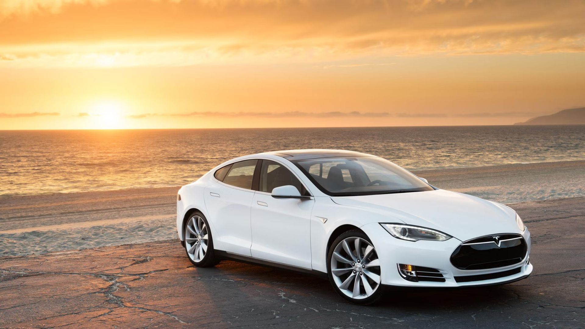 Front three-quarter view of a white 2015 Tesla Model S parked on a beach as the sun sets.