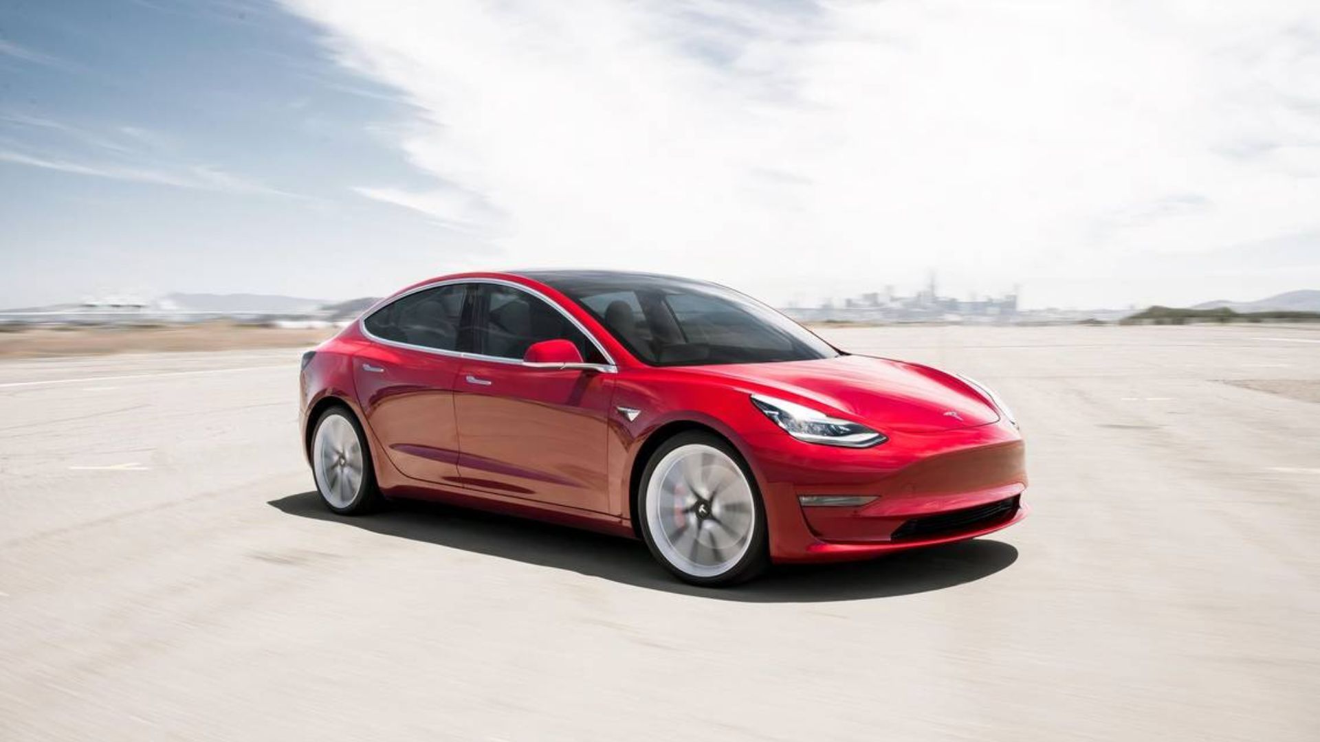 Full view of a red 2018 Tesla Model 3 driving on an airfield.