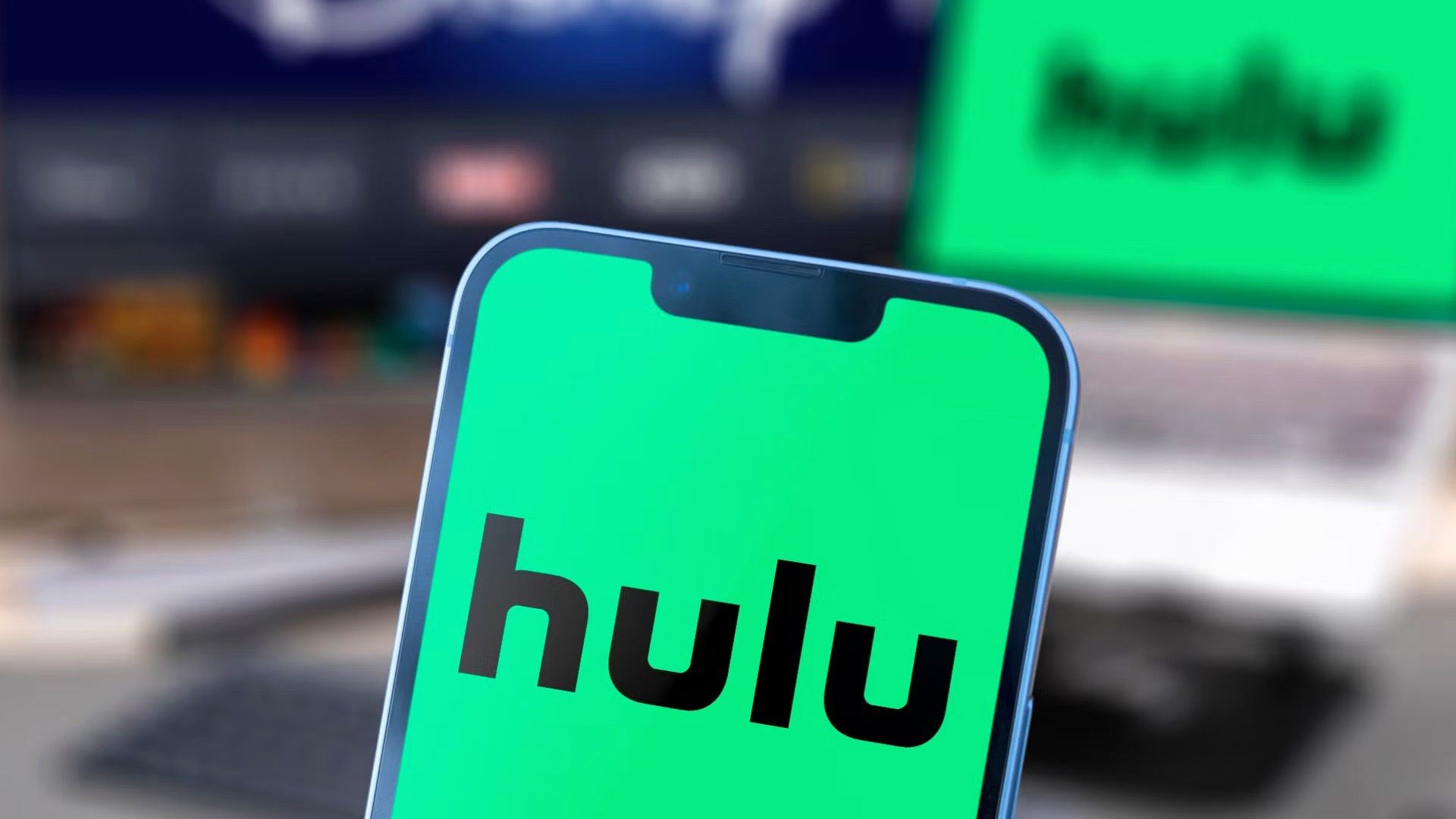 a phone with the hulu logo and a monitor and laptop with the disney plus and hulu logo blurred in the background
