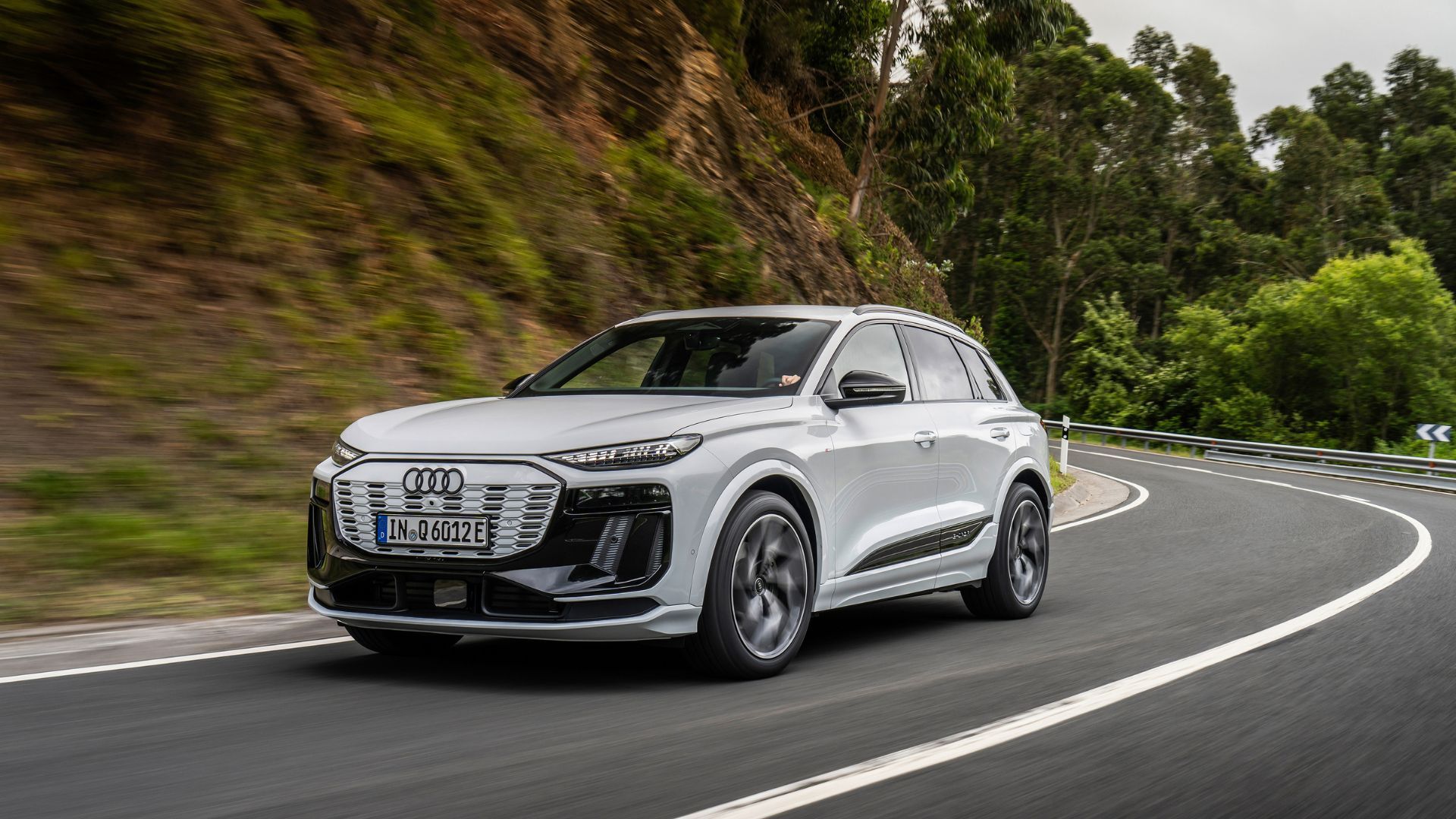 Full view of a a white Audi Q6 e-tron driving on a winding country road.