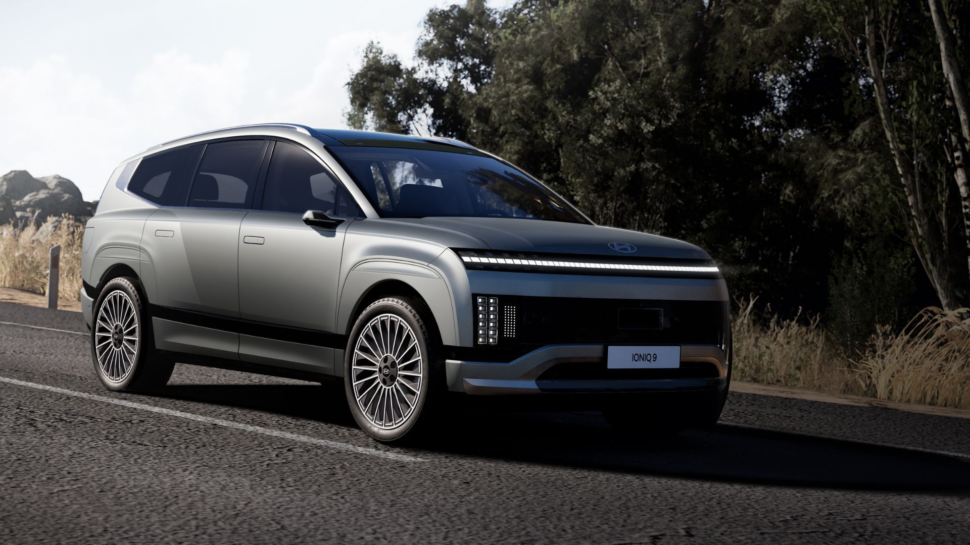 Full view of a gray 2026 Hyundai IONIQ 9 driving on a country road with trees in the background.