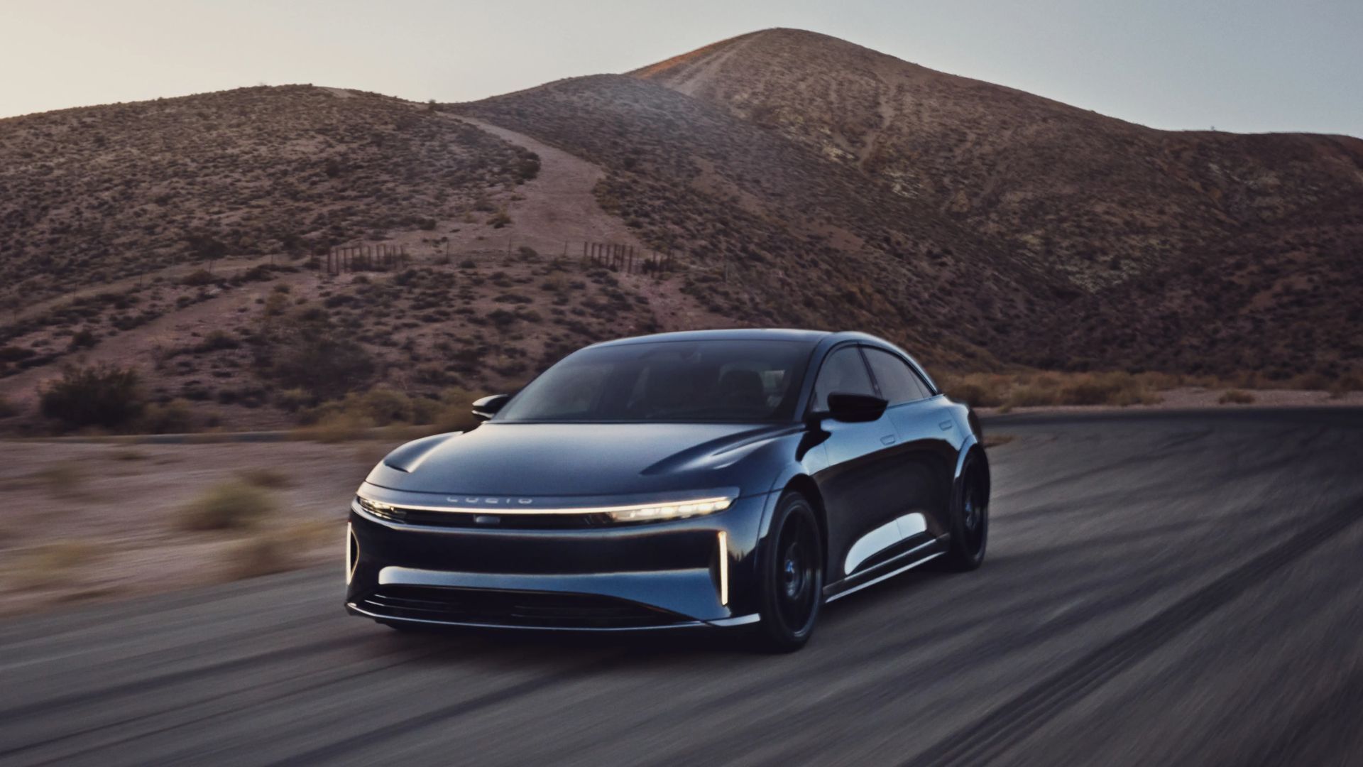 Full view of a blue Lucid Air Sapphire driuving on a country road with a mountain in the background.