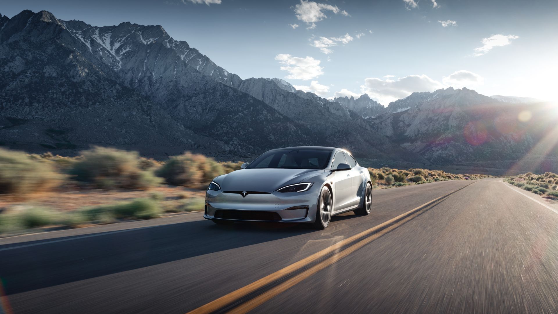 Front three-quarter view of a silver Tesla Model S driving on a country road the mountains.