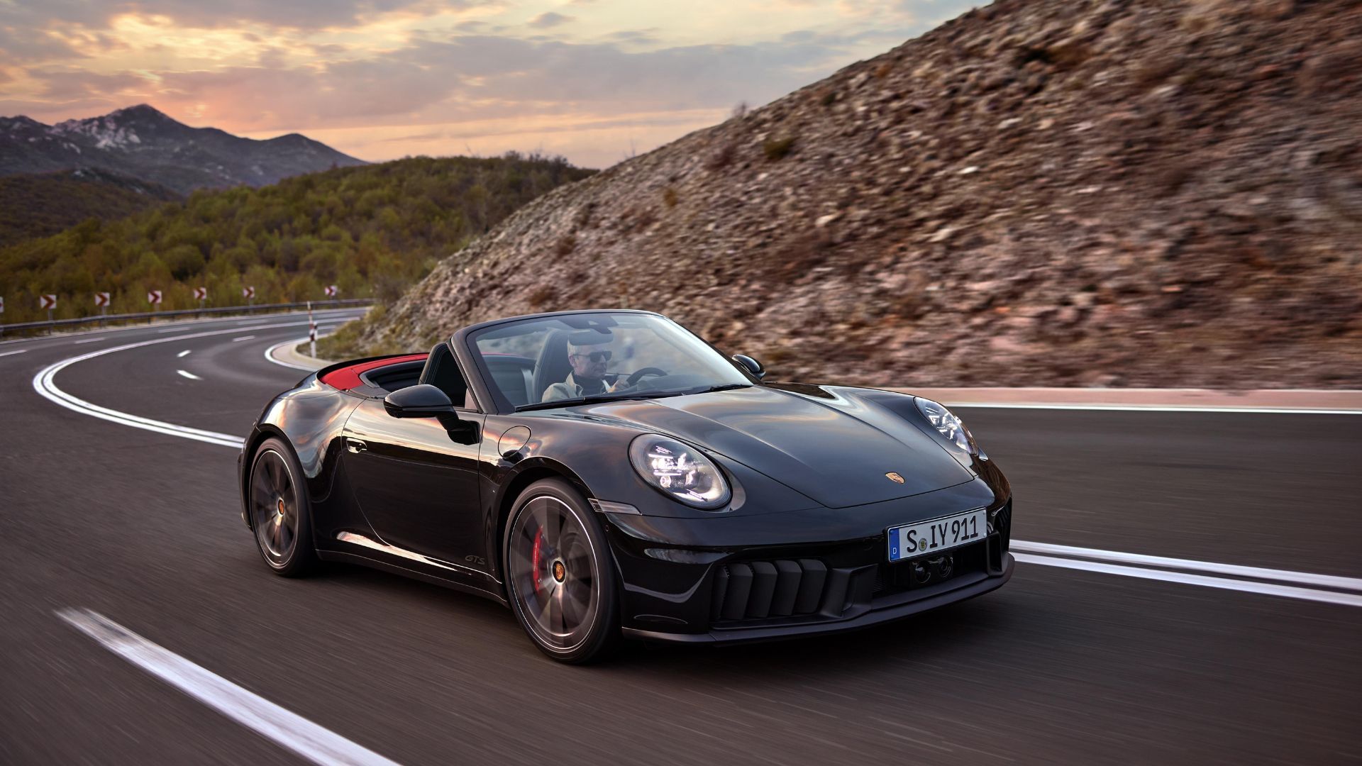 Full view of a black 2024 Porsche 911 Carrera GTS Cabriolet Hybrid driving on a winding country road with mountains in the background.