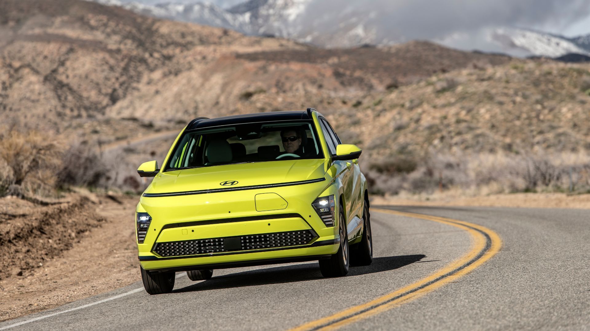 Dynamic front 3/4 shot of a yellow 2025 Hyundai Kona Electric driving on a winding country road.