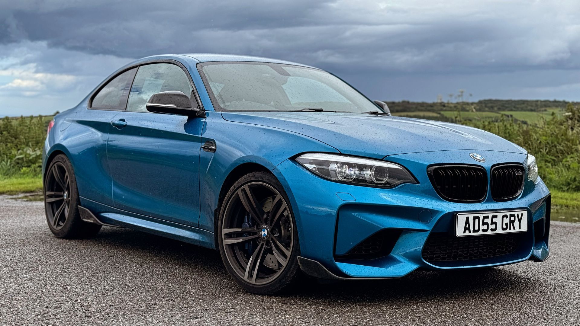 Static front 3/4 shot of a blue 2018 BMW M2 with fields in the background.