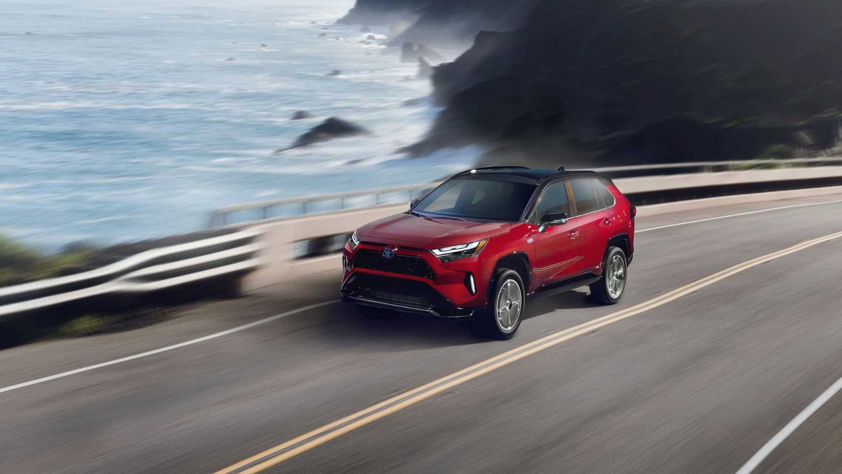 Dynamic front 3/4 shot of a red 2024 Toyota RAV4 Prime XSE driving on a road running beside the ocean.