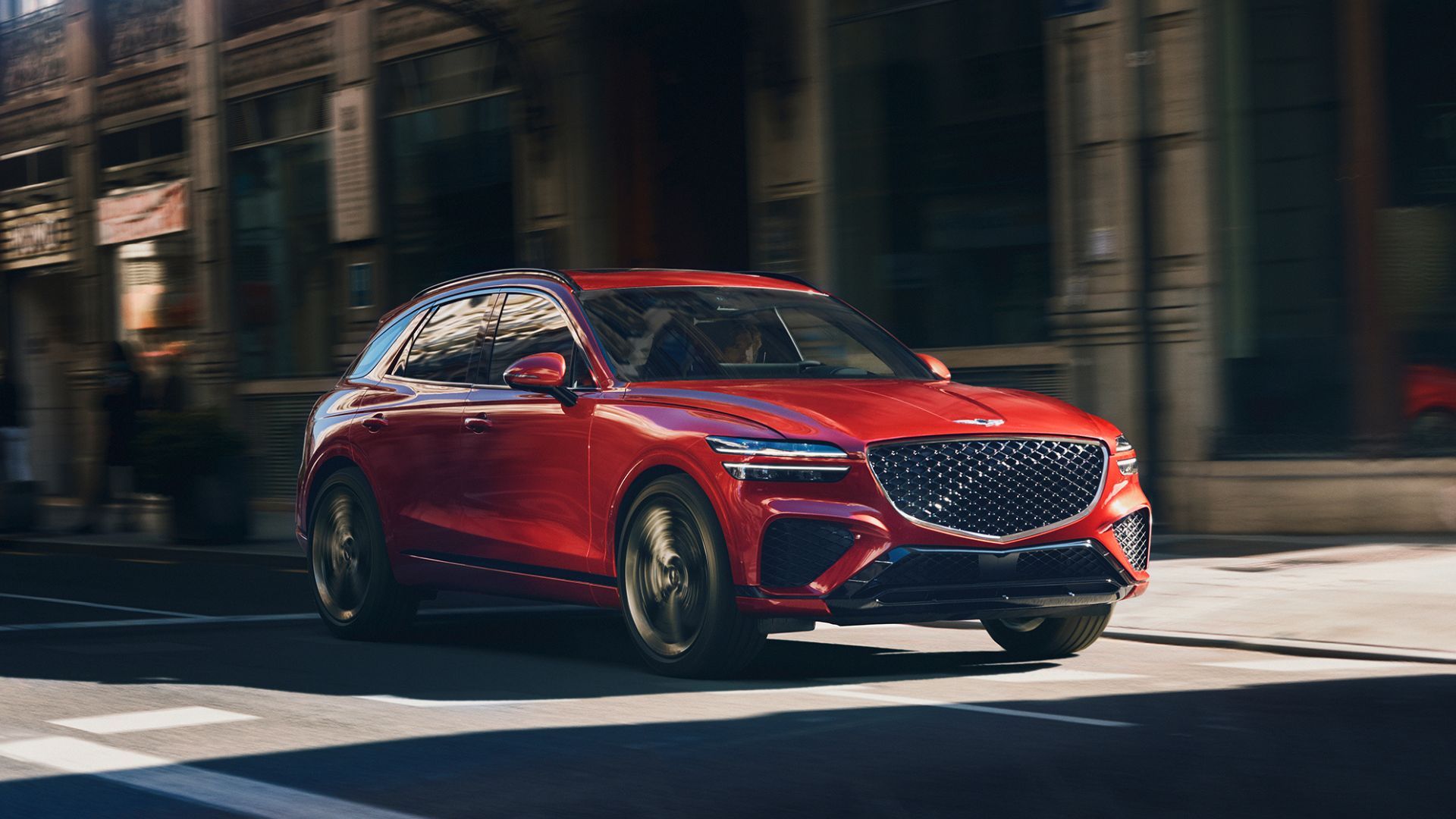 Dynamic front 3/4 shot of a red Genesis GV70 driving through a city.