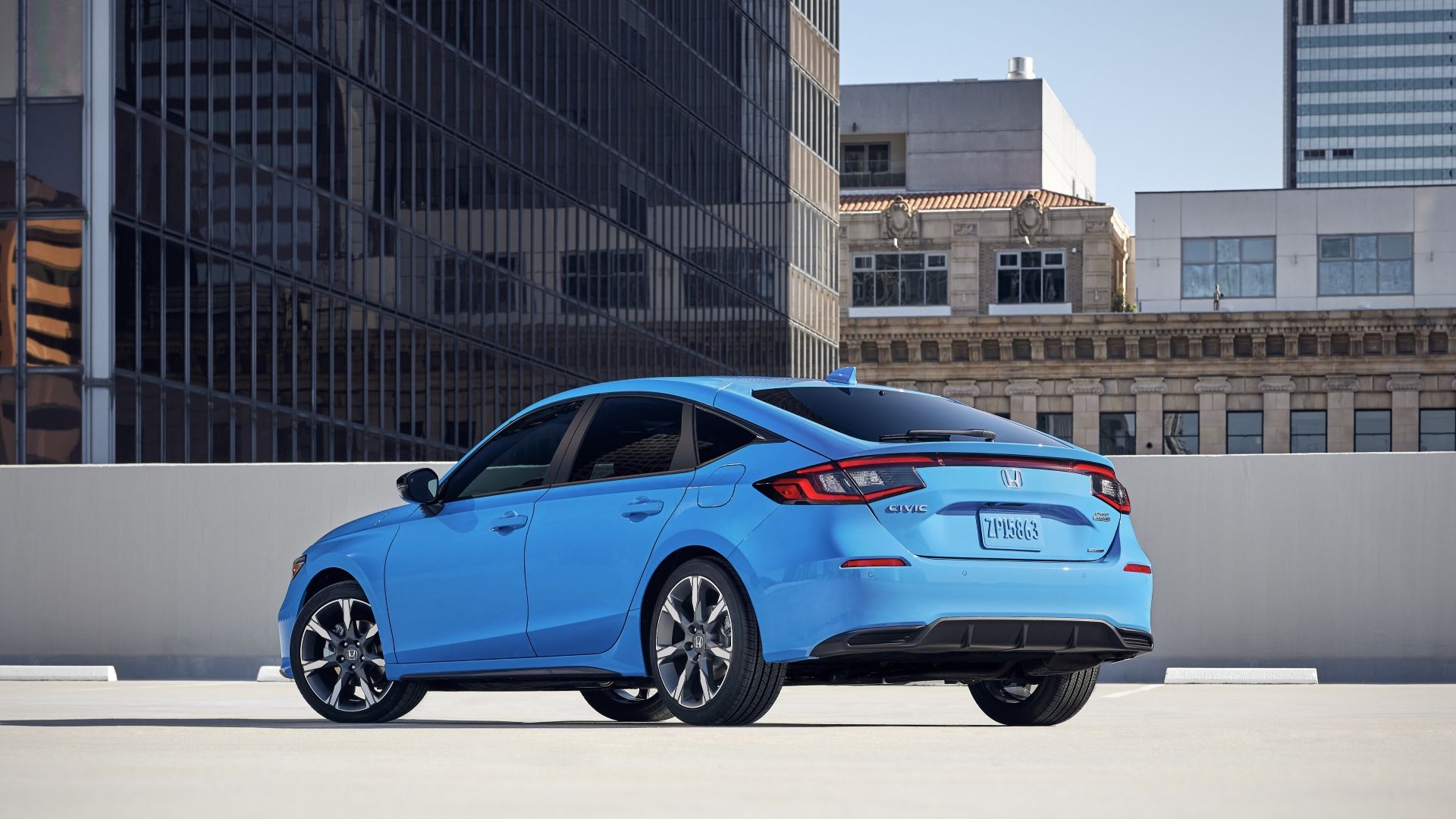 Static rear 3/4 shot of a blue 2025 Honda Civic Hatchback parked on a rooftop with tall buildings in the background.