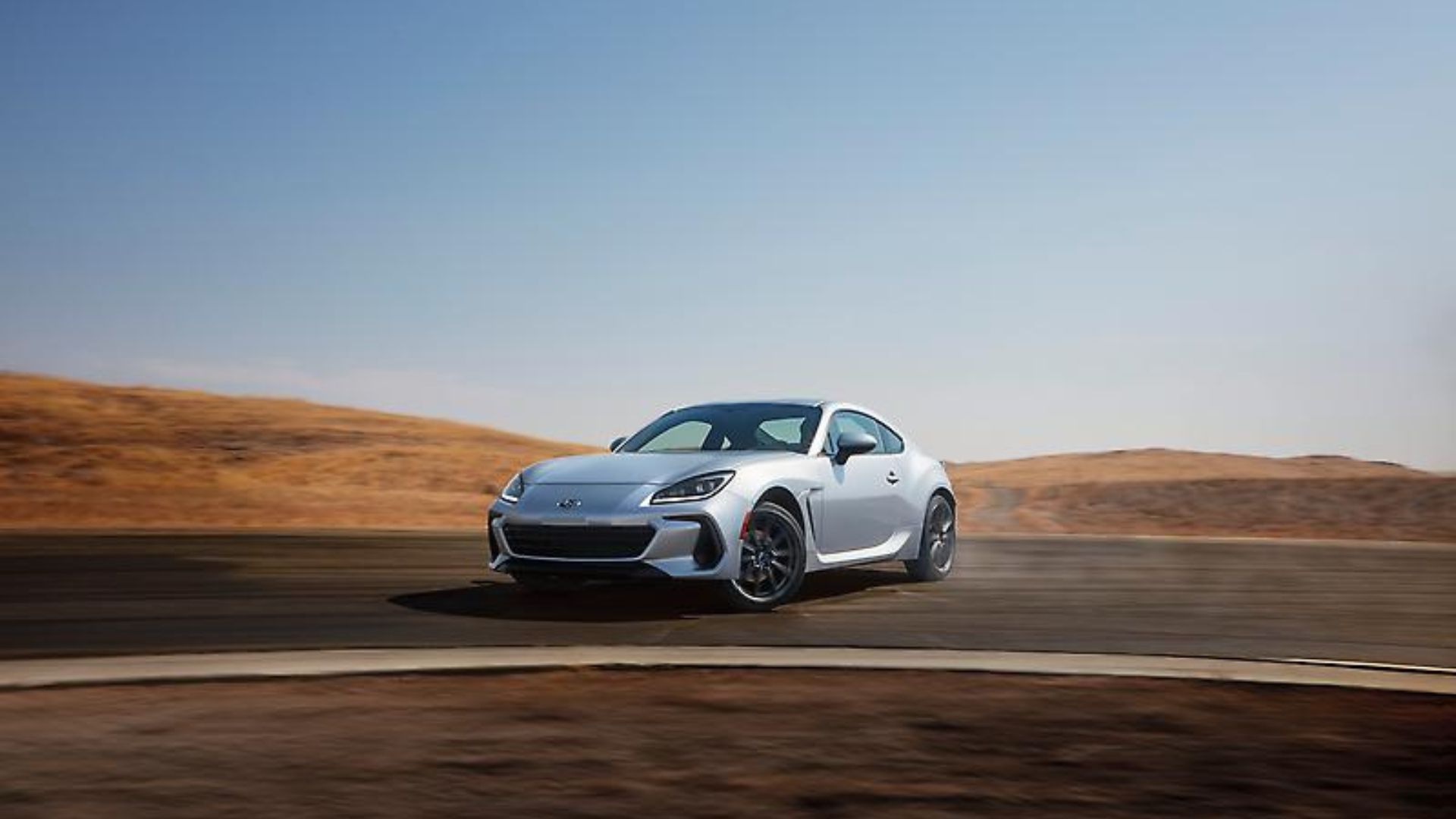 Dynamic front 3/4 shot of a silver 2024 Subaru BRZ drifting around a corner on a racetrack.