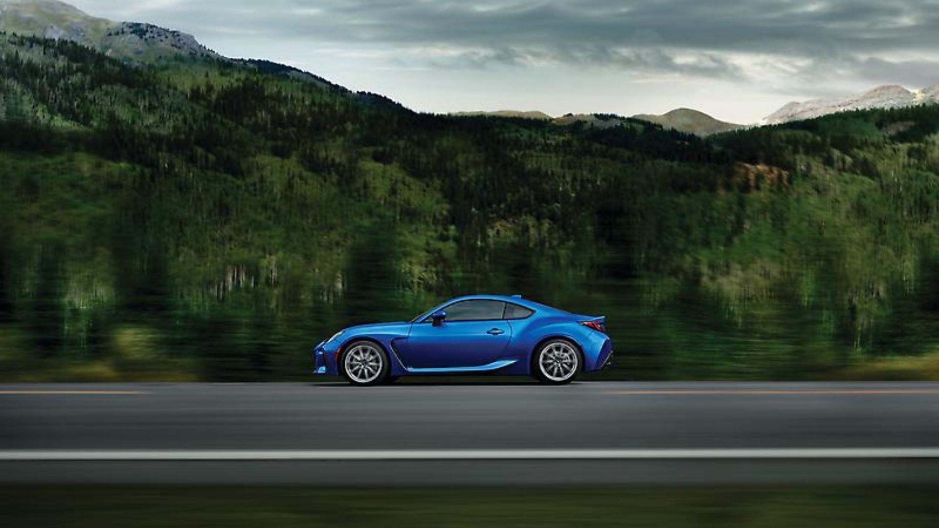Dynamic side profile shot of a blue 2024 Subaru BRZ driving on a country road with mountains and trees in the background.