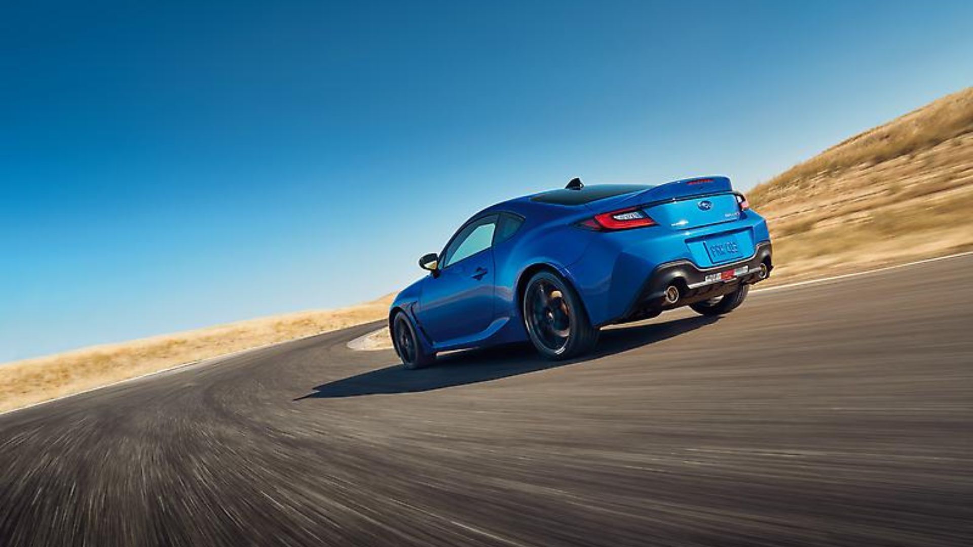 Dynamic rear 3/4 shot of a blue 2024 Subaru BRZ tS driving round on a corner on a country road.