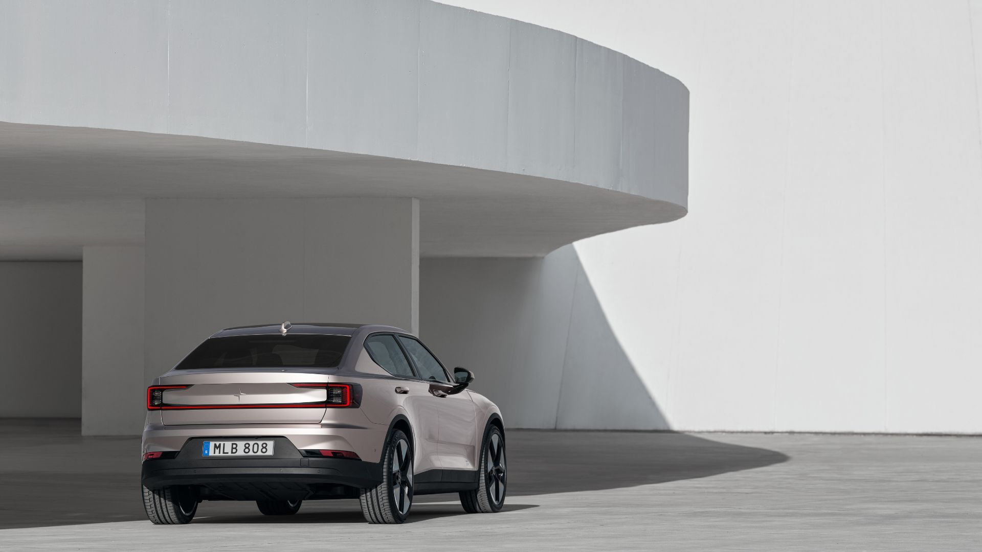 Static rear 3/4 shot of a gold 2023 Polestar 2 parked outside of a modern building.