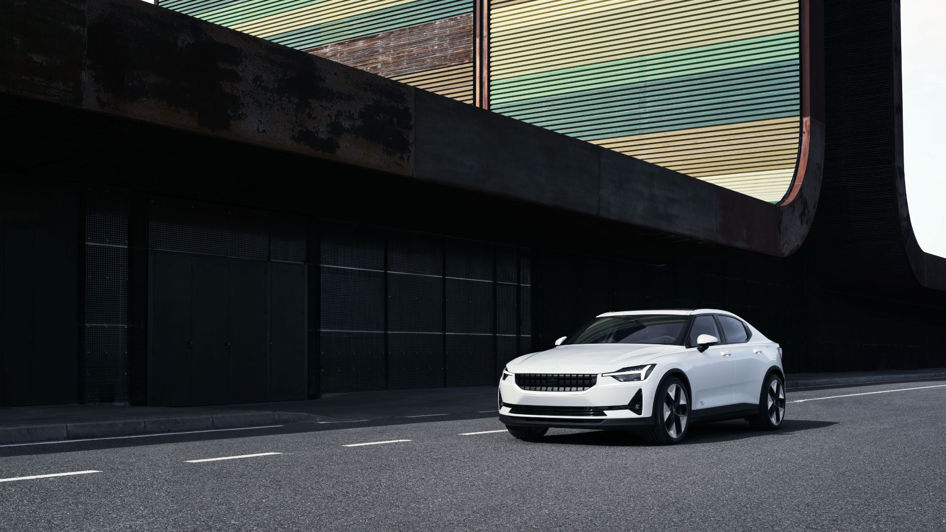 Dynamic front 3/4 shot of a white 2023 Polestar 2 driving through a city with a modern building in the background.