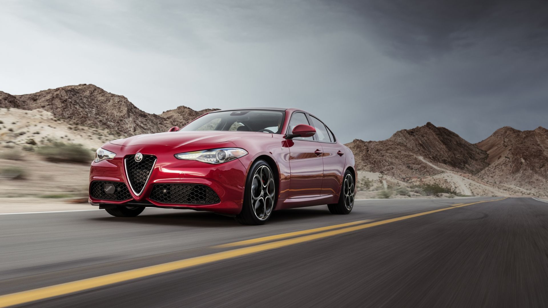 Dynamic front 3/4 shot of a red 2022 Alfa Romeo Giulia drivingh ion a country road with mountains in the background.