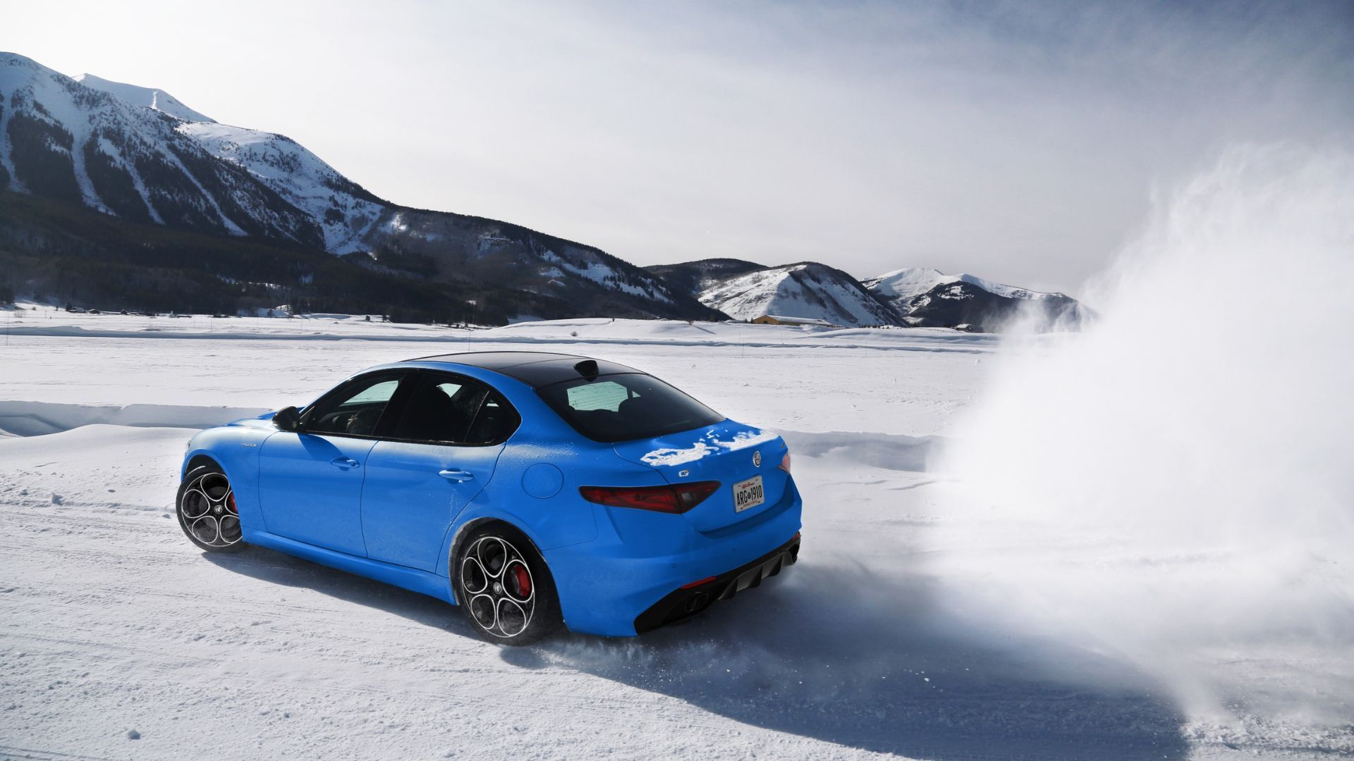 Dynamic rear 3/4 shot of a blue 2022 Alfa Romeo Giulia thrashing around in the snow with mountains in the background.