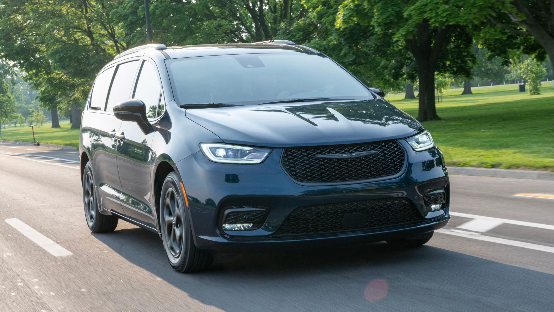 Dynamic front 3/4 shot of a black 2025 Chrysler Pacifica Plug-in Hybrid driving through a city with grass and trees in the background.