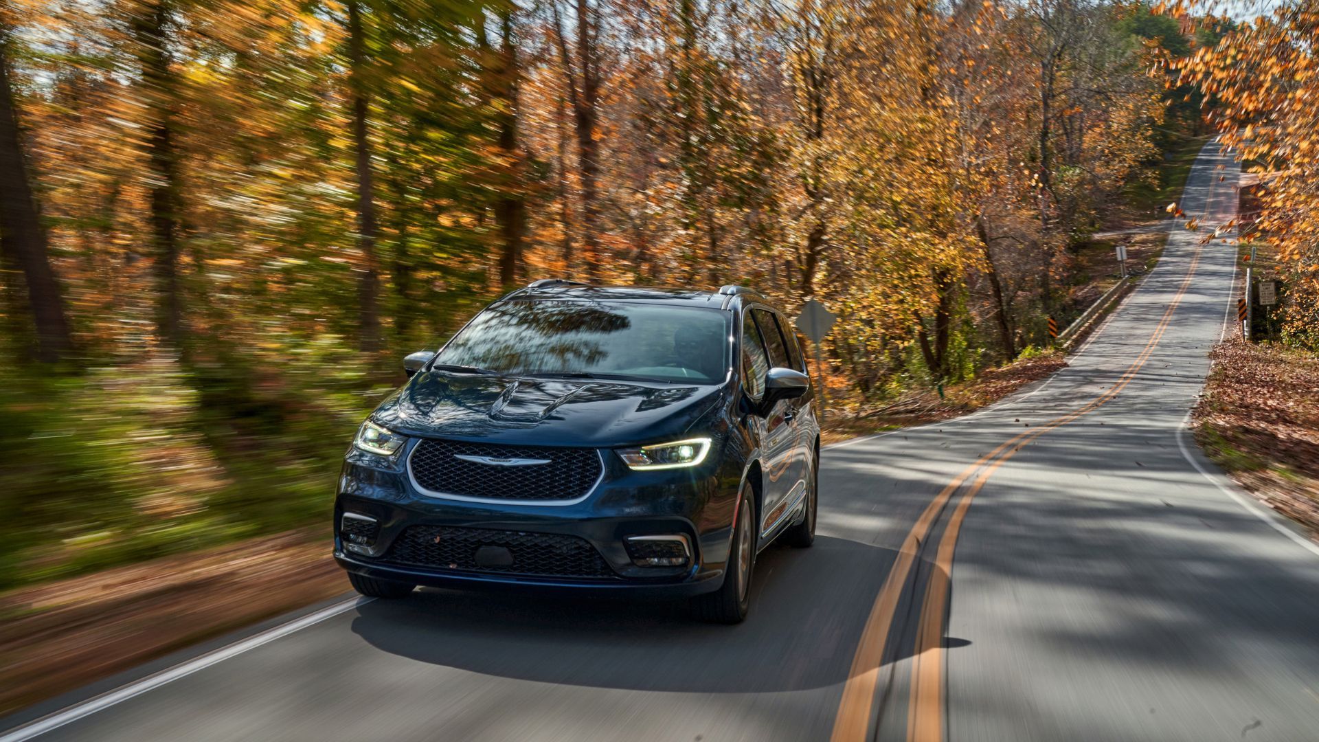 Dynamic front 3/4 shot of a dark blue 2025 Chrysler Pacifica Plug-in Hybrid Pinnacle driving on a country road lined by trees.